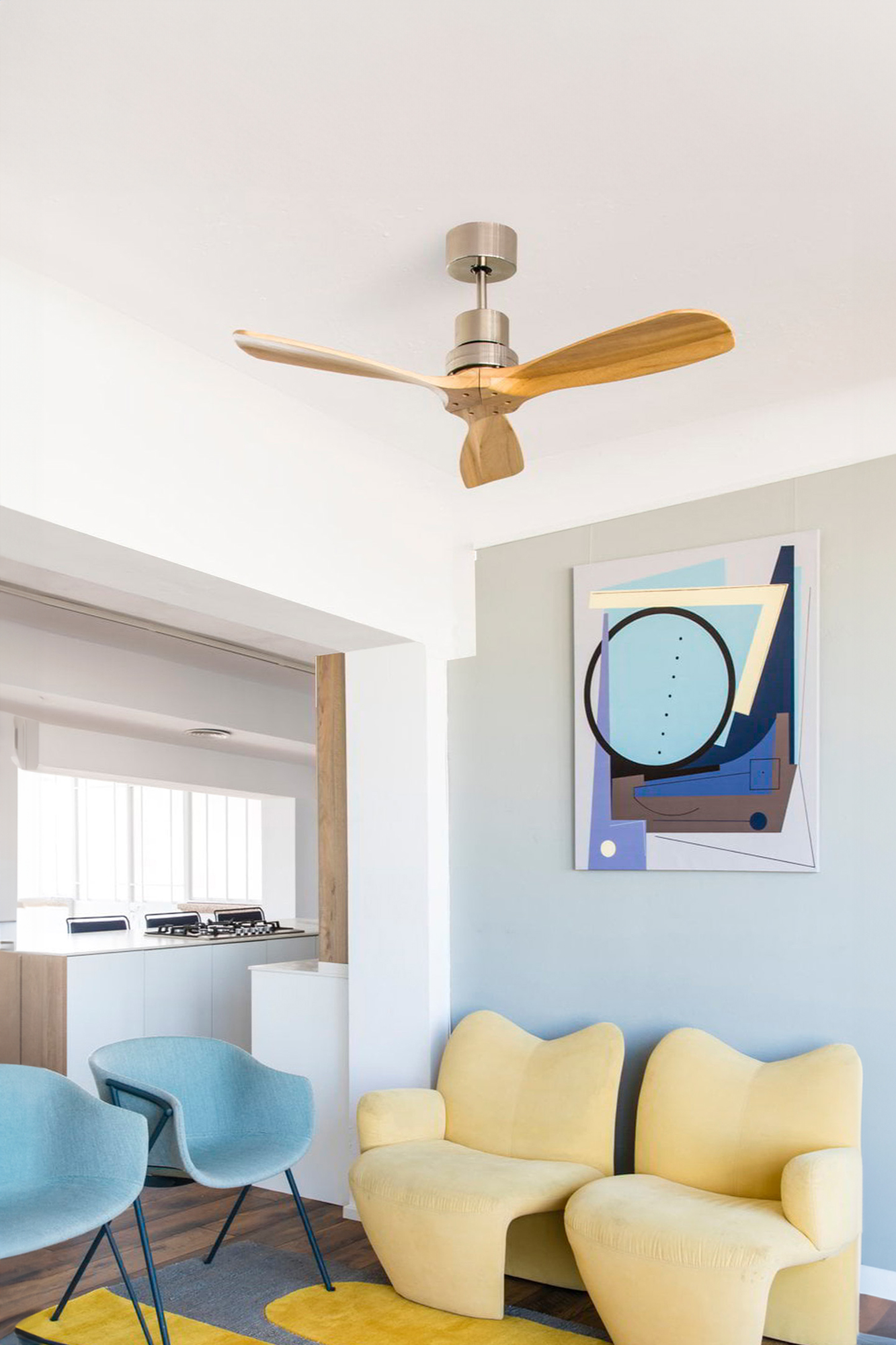 Stanza con ventilatore a soffitto, sedie e opere d'arte. Arredamento giallo e blu con tappeto grigio e soffitto bianco. Cucina sullo sfondo.