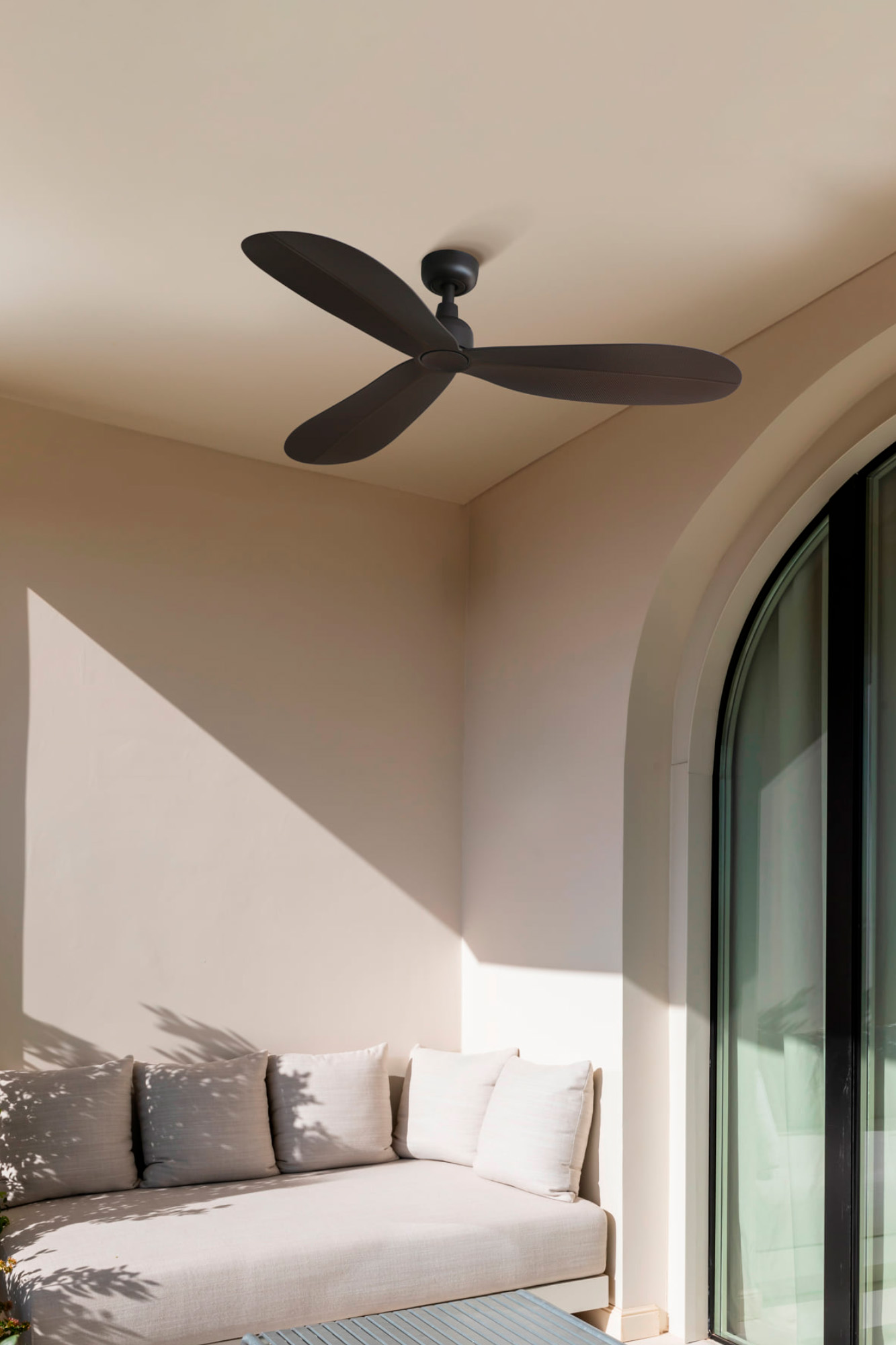 Ein Deckenventilator hängt über einem gepolsterten Sitzbereich. Der Raum hat eine beige Wand mit Schatten und eine gewölbte Glastür.