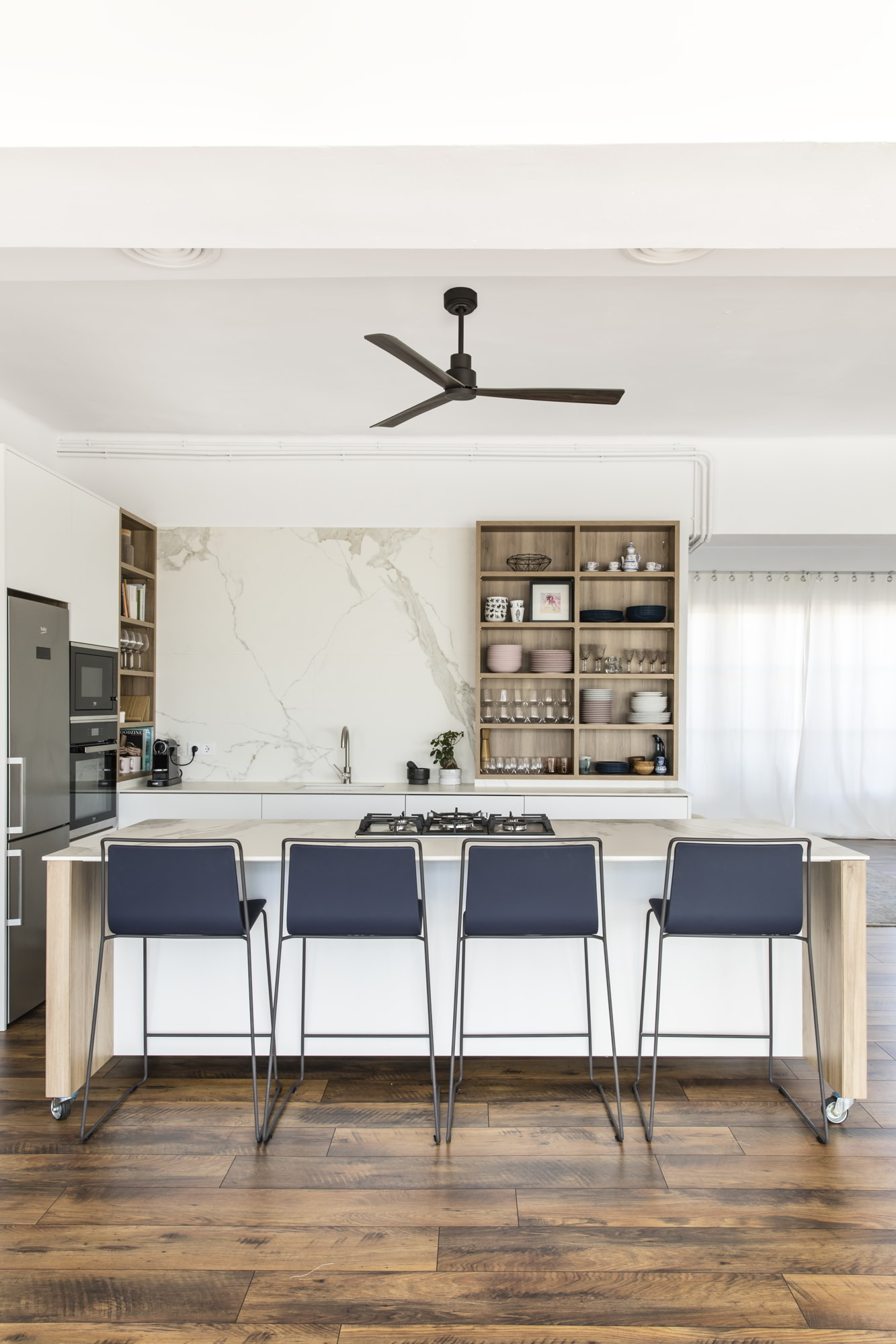 Cucina moderna con isola, quattro sgabelli blu, pavimento in legno, armadi bianchi, paraschizzi in marmo e ventilatore a soffitto.