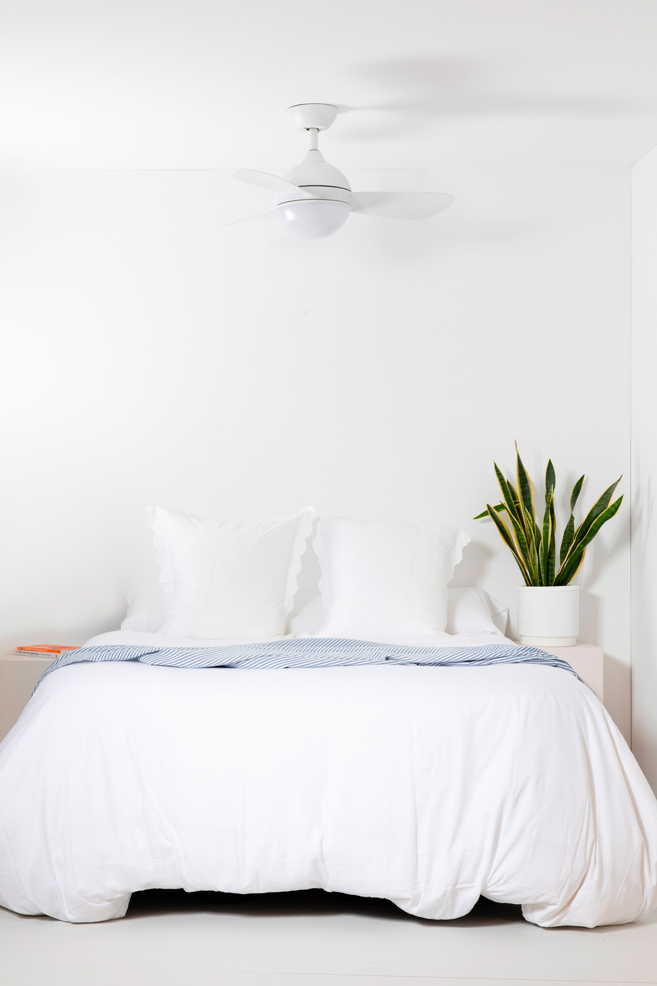 Schlafzimmer mit weißem Bett, Kissen und Bettwäsche. Eine Pflanze steht auf einem Nachttisch. Über dem Bett befindet sich ein Deckenventilator.