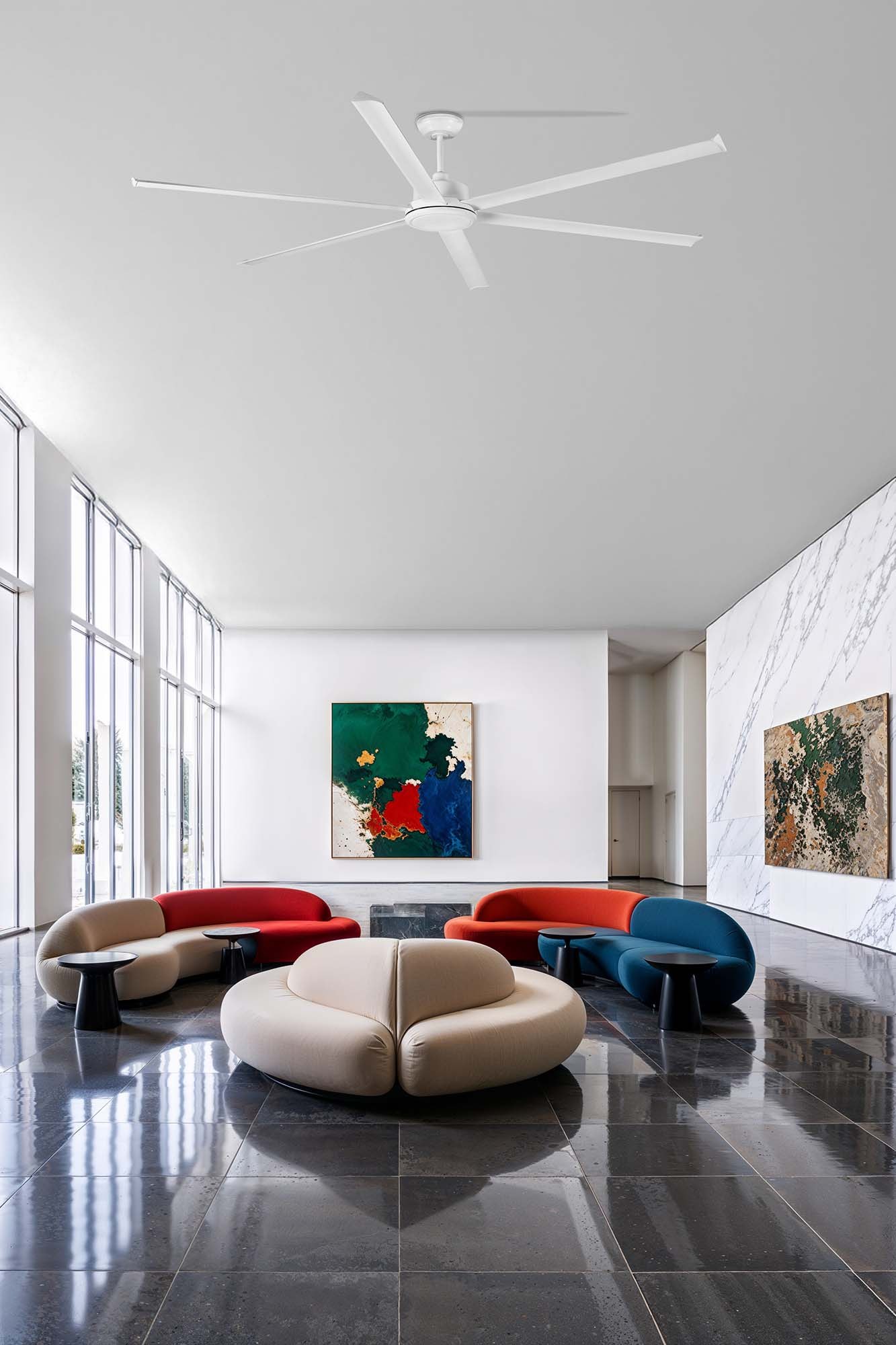 Un interno moderno presenta divani colorati, opere d'arte e un grande ventilatore a soffitto. Il pavimento è lucido e si vede un muro di marmo.