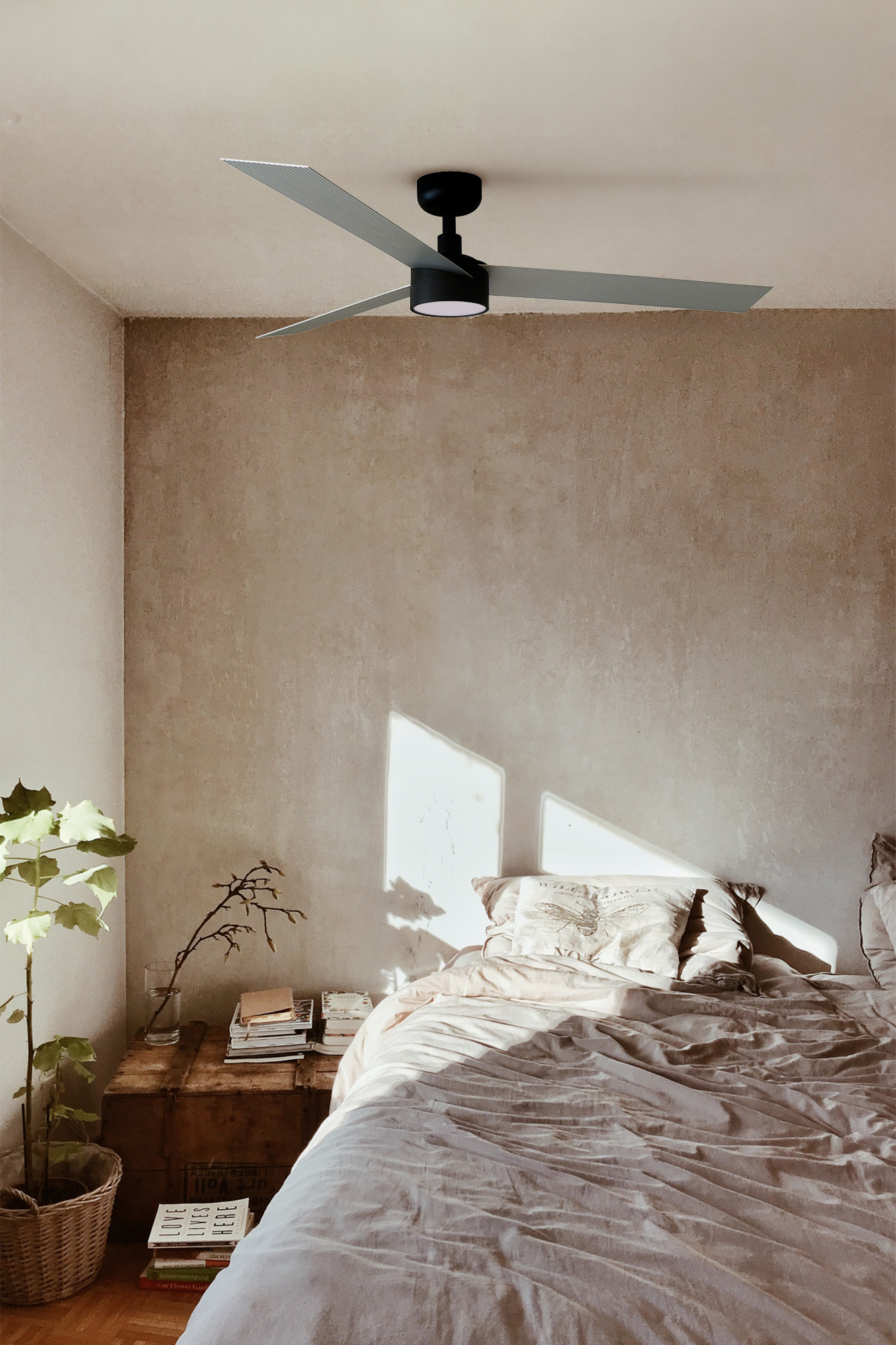 Schlafzimmer mit Bett, Ventilator und Holzkiste. Licht scheint herein. Pflanze und B ücher stehen neben dem Bett.