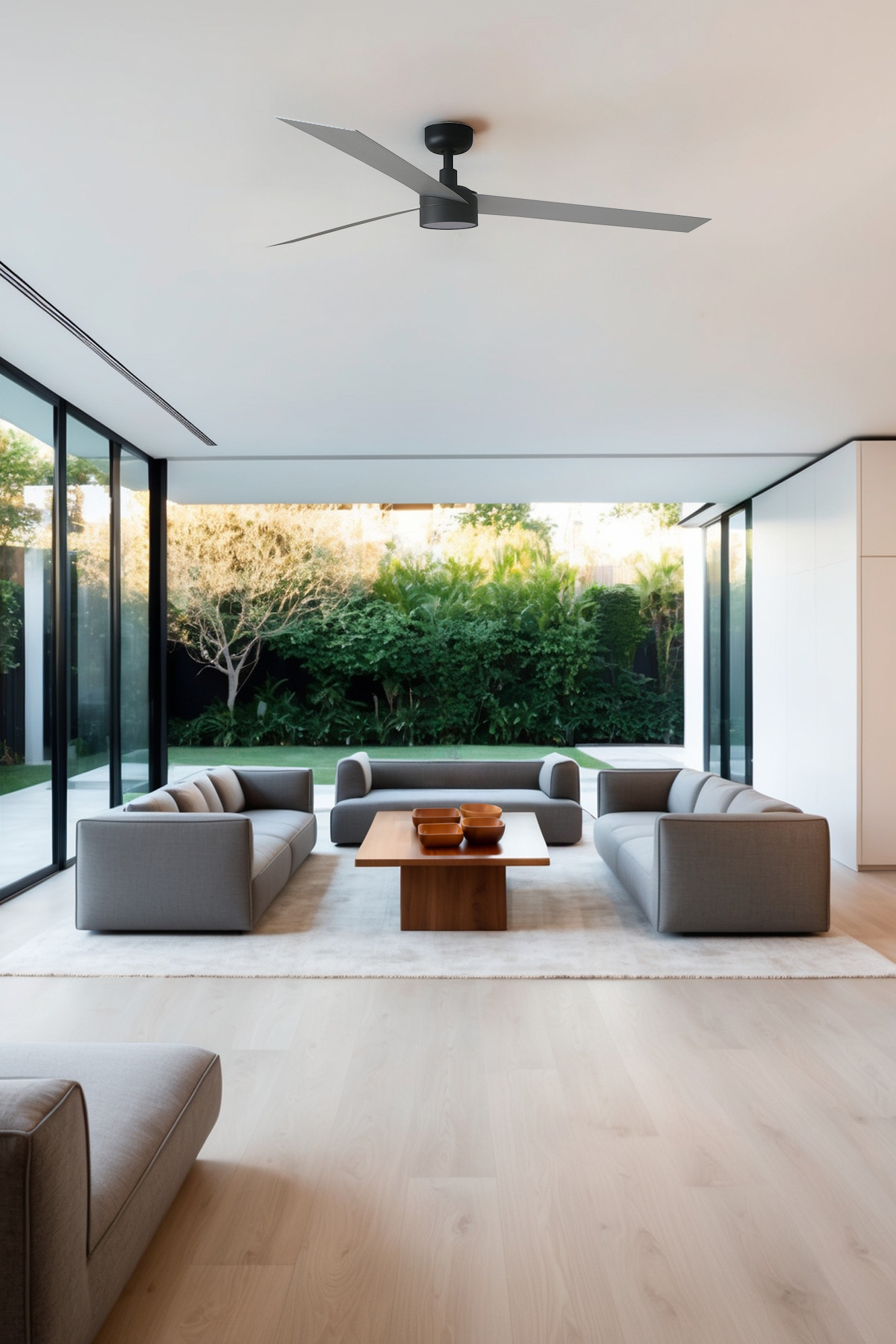 Modernes Wohnzimmer mit grauen Sofas, Holztisch und großen Fenstern mit Blick auf einen grünen Garten. Ein Deckenventilator ist sichtbar.