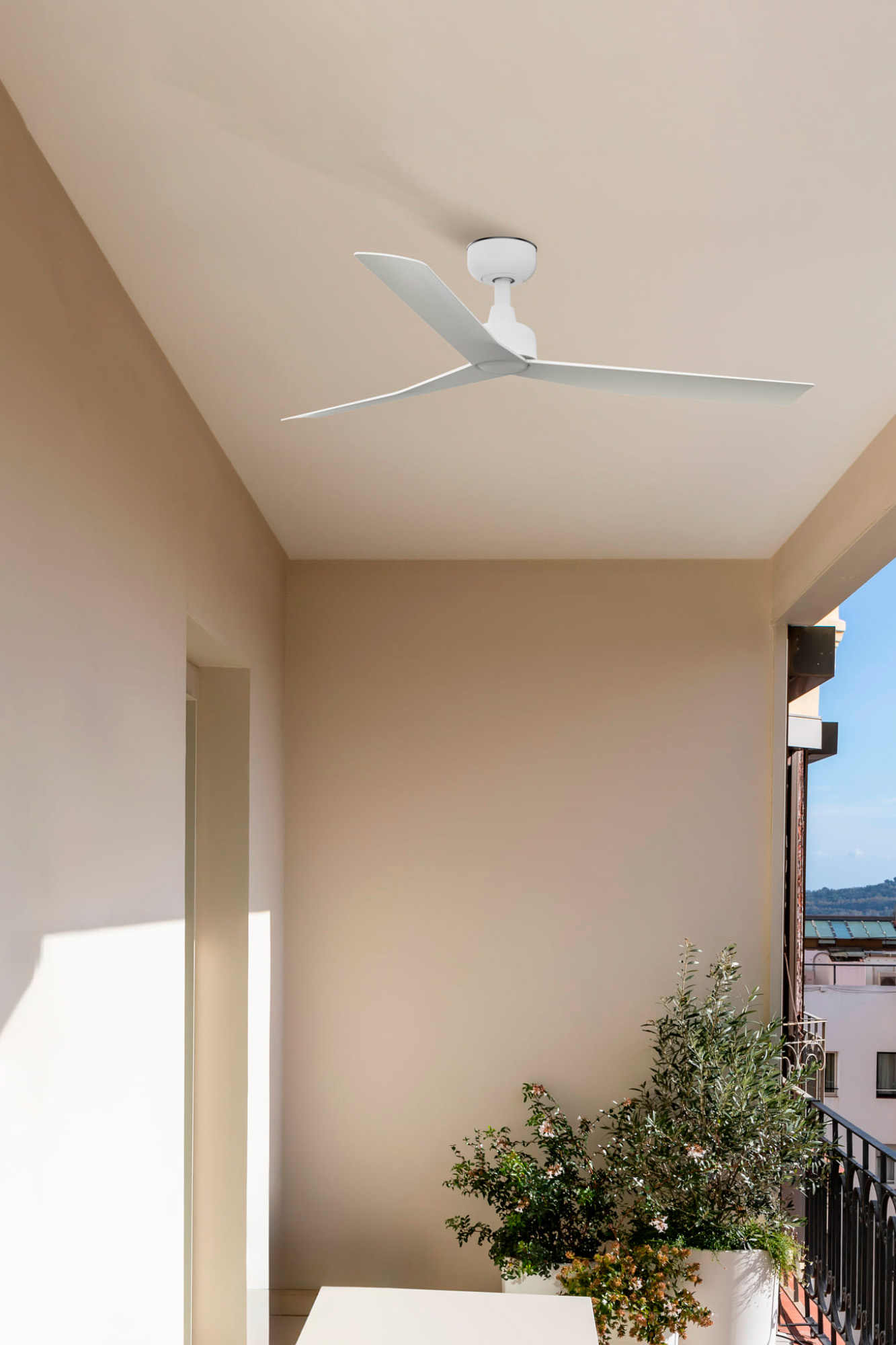 Un ventilatore da soffitto bianco è appeso sopra un balcone. Un vaso di fiori con vegetazione è sulla destra. Muri beige e cielo blu sono visibili.