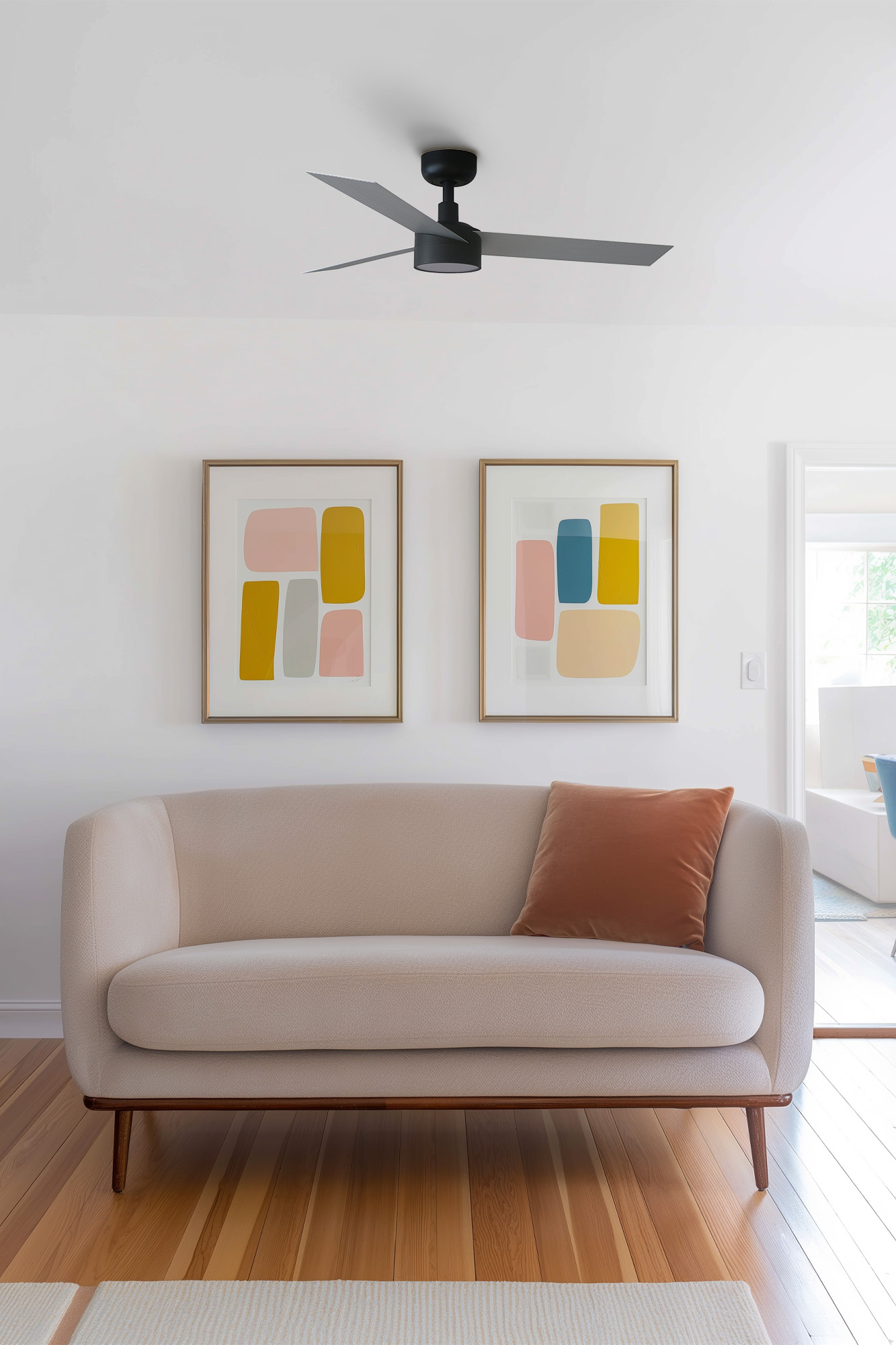 Wohnzimmerszene mit beigem Sofa, Kunstdrucken und Deckenventilator. Oranges Kissen auf dem Sofa, Holzboden und weiße Wände.