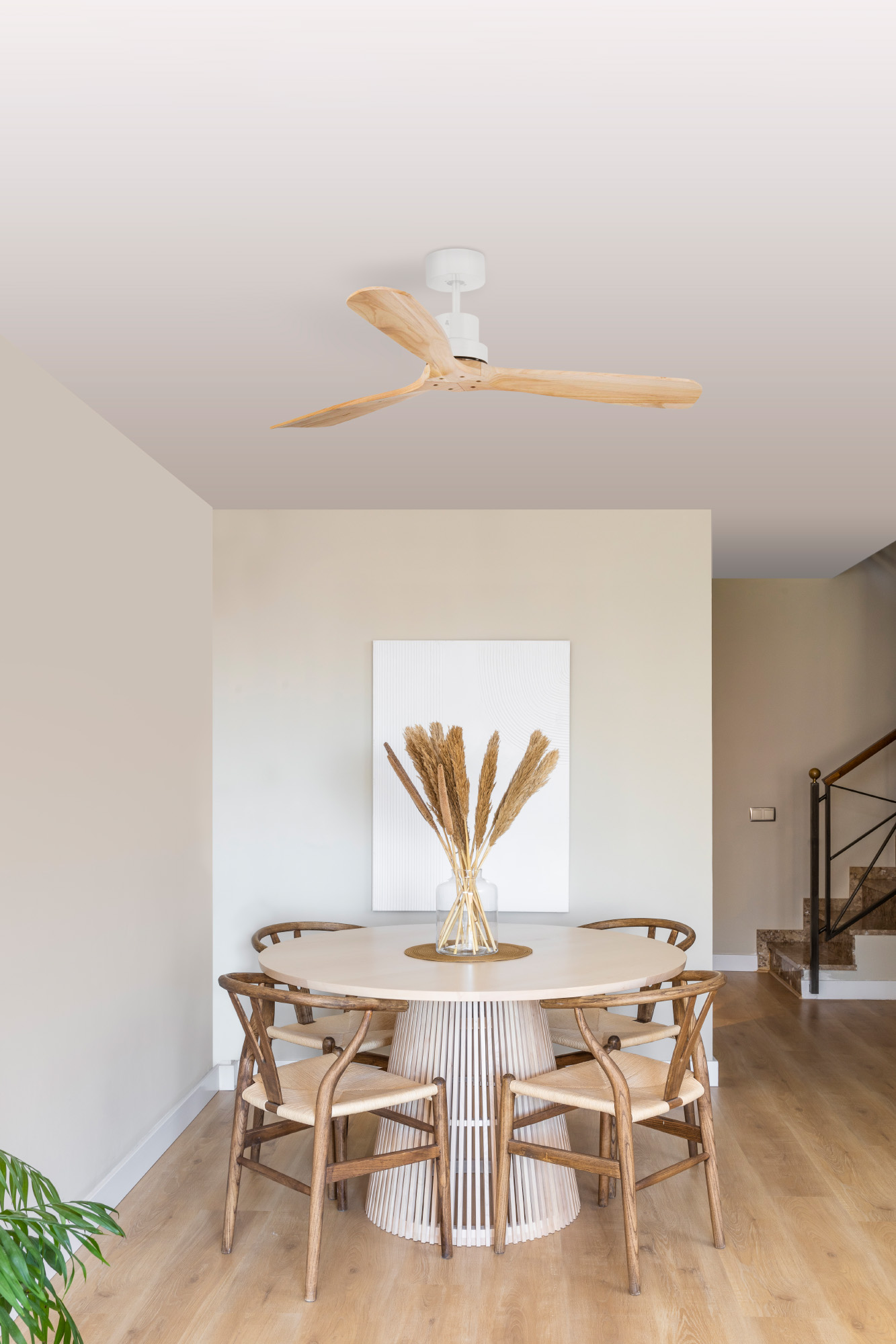 Ein Esszimmer mit rundem Tisch, Stühlen, Deckenventilator und einer Vase mit getrockneten Pflanzen. Holzboden und Treppe im Hintergrund.