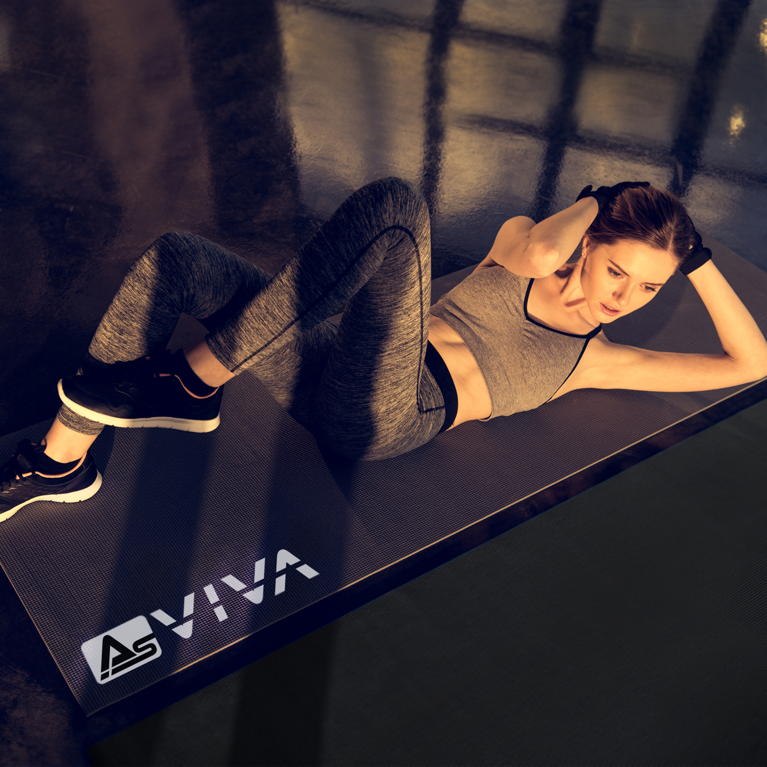 Eine Frau in Trainingskleidung macht Sit-ups auf einer Matte. Sie liegt auf einem Gymnastikboden und hat die Hände hinter dem Kopf. Das Logo lautet AS VIVA.