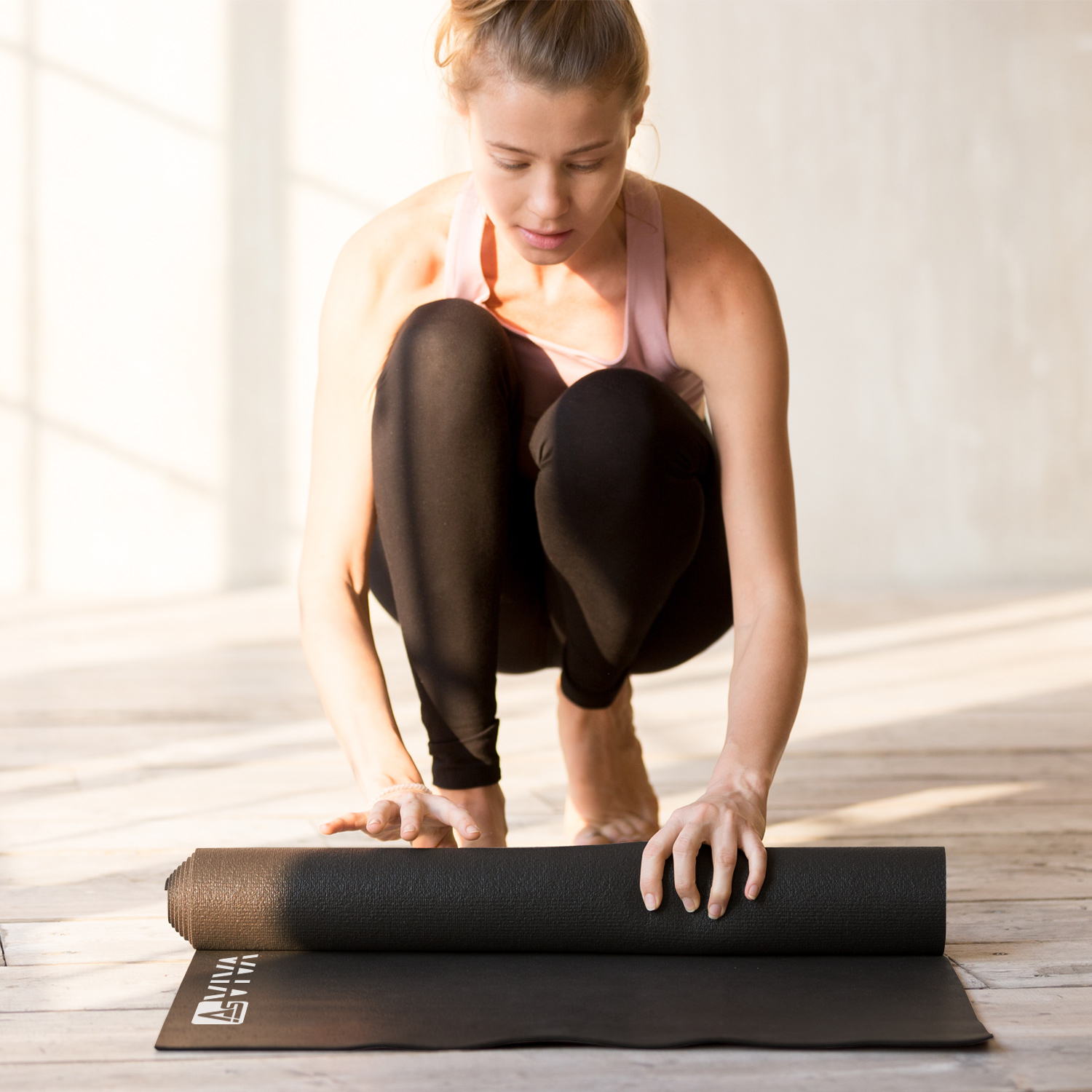 Eine Frau in Sportkleidung rollt eine Yogamatte aus. Sie hockt sich hin und bereitet sich auf das Training auf einem Holzboden vor.