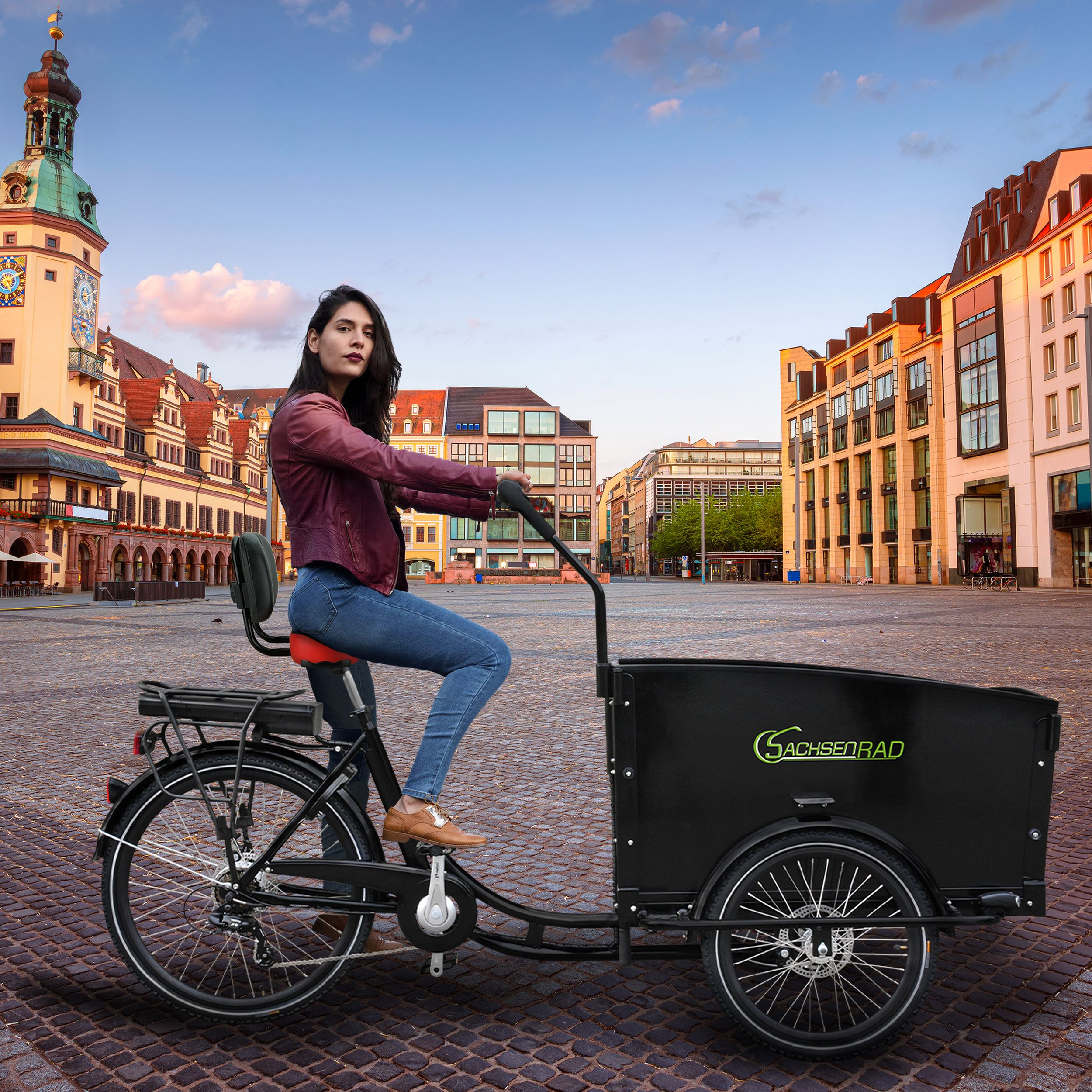 Eine Frau fährt mit einem schwarzen Lastenrad auf einem Stadtplatz. Gebäude und ein Glockenturm befinden sich im Hintergrund.