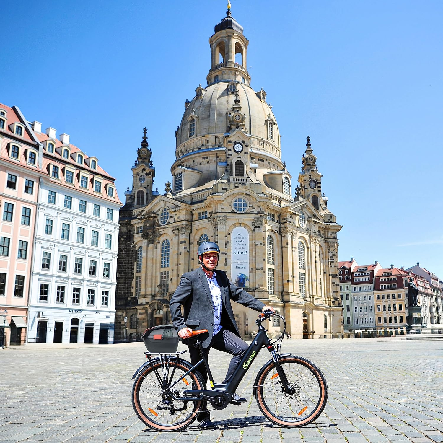Ein Mann in Anzug und Helm posiert auf einem Fahrrad vor einer großen Kathedrale und anderen Gebäuden an einem sonnigen Tag.