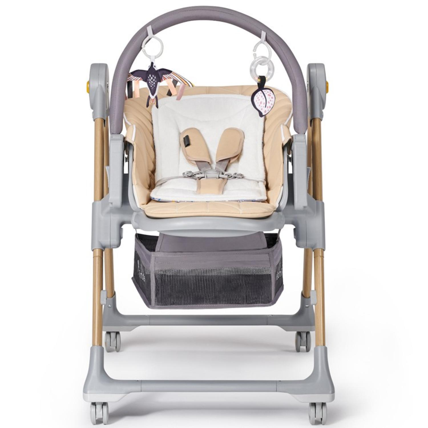 Ein beiger und weißer Babyhochstuhl mit Spielzeug, das von oben herabhängt. Er hat ein graues Gestell und Holzbeine, einen grauen Korb und Räder.