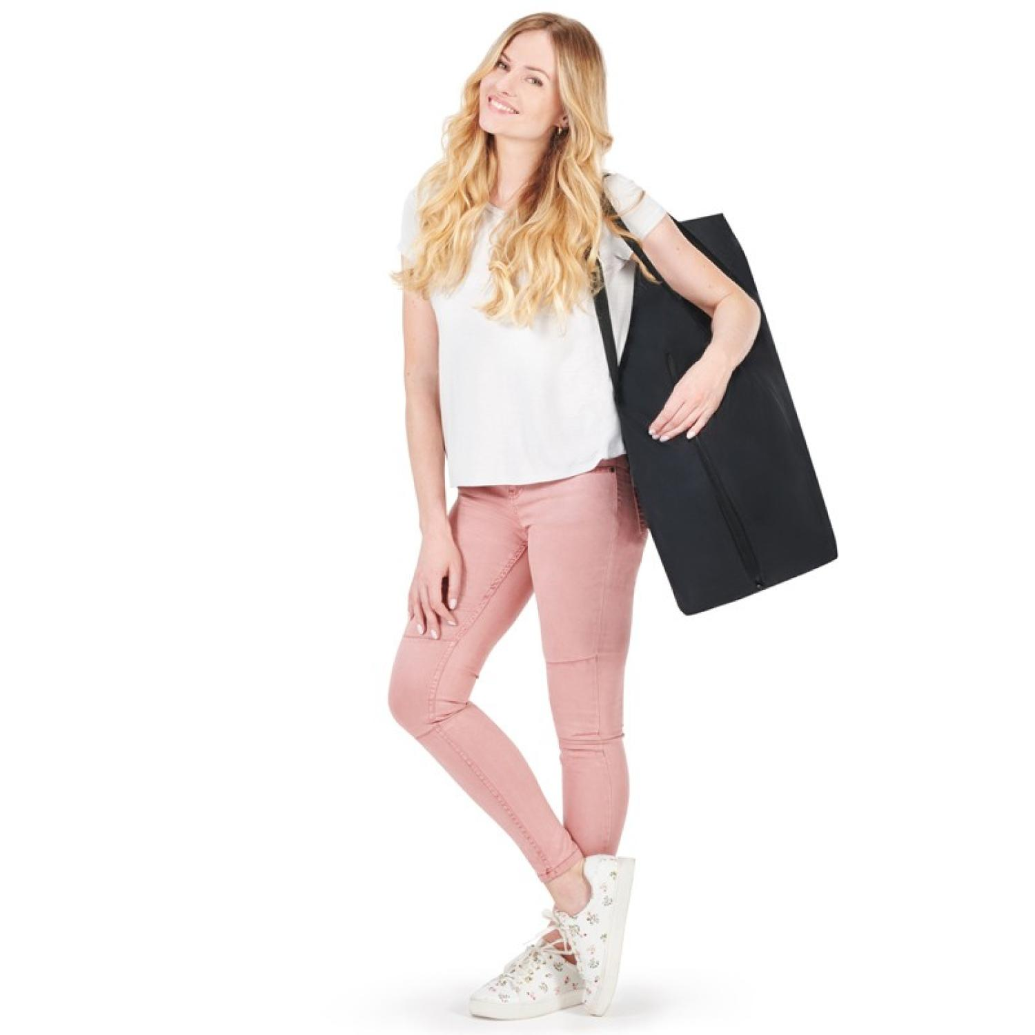 Blonde Frau in weißem T-Shirt und rosa Hose hält eine schwarze Tasche und lächelt. Sie trägt weiße Turnschuhe. Weißer Hintergrund.