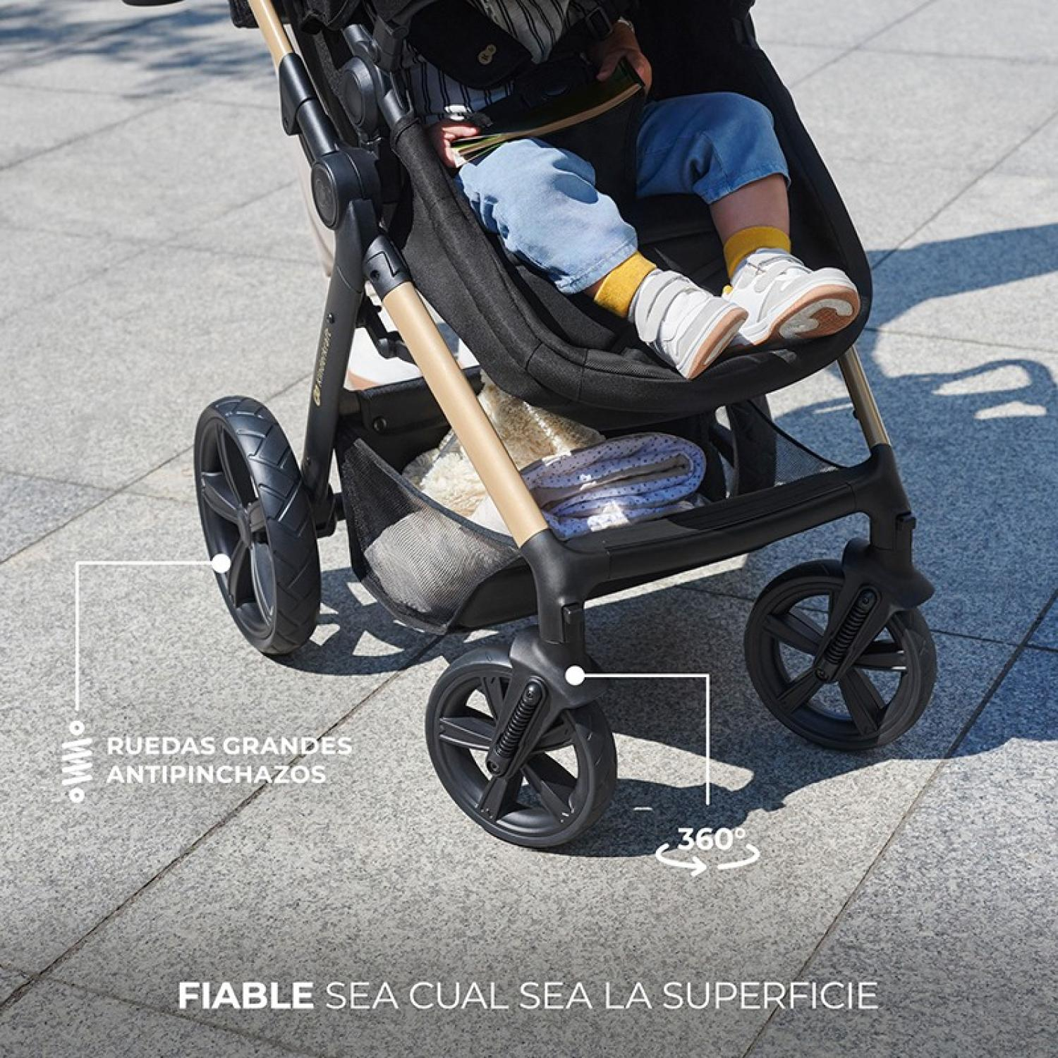 Ein Baby sitzt in einem schwarzen Kinderwagen mit großen Rädern. Der Kinderwagen hat einen beigefarbenen Rahmen und steht auf einer gepflasterten Oberfläche.