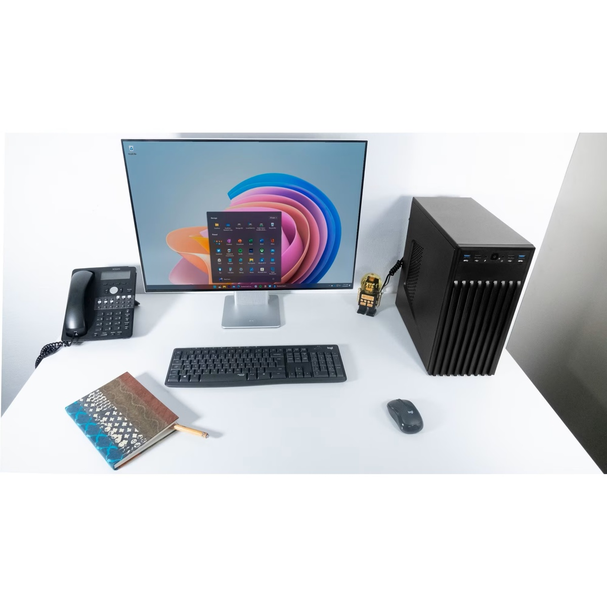 Biurko z komputerem, monitorem, klawiaturą, myszą i czarnym telefonem. Na biurku znajduje się również notatnik i długopis.