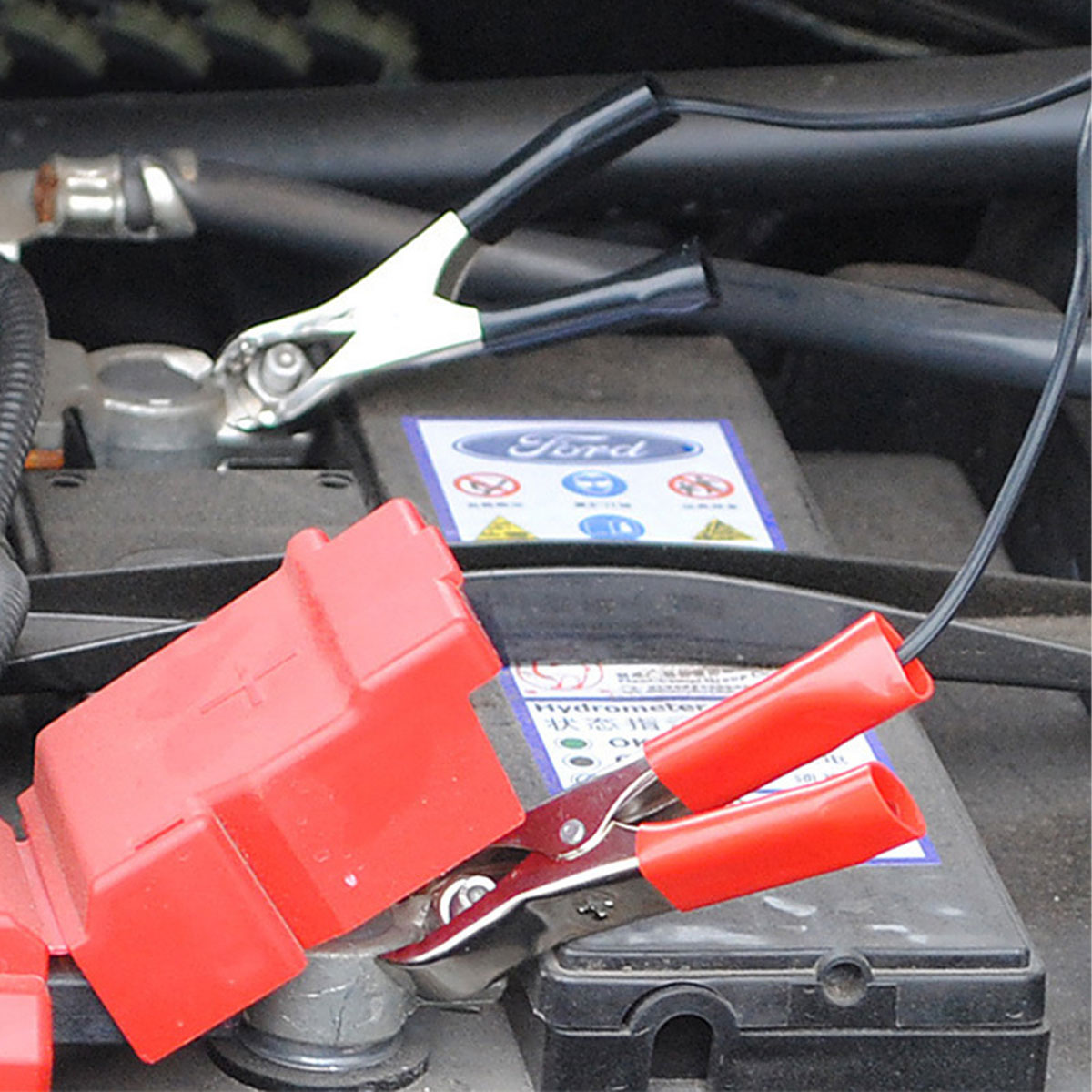 Autobatterie mit angeschlossenen Überbrückungskabeln. Rote und schwarze Klemmen sind an den Klemmen befestigt. Ein Ford-Logo ist sichtbar.