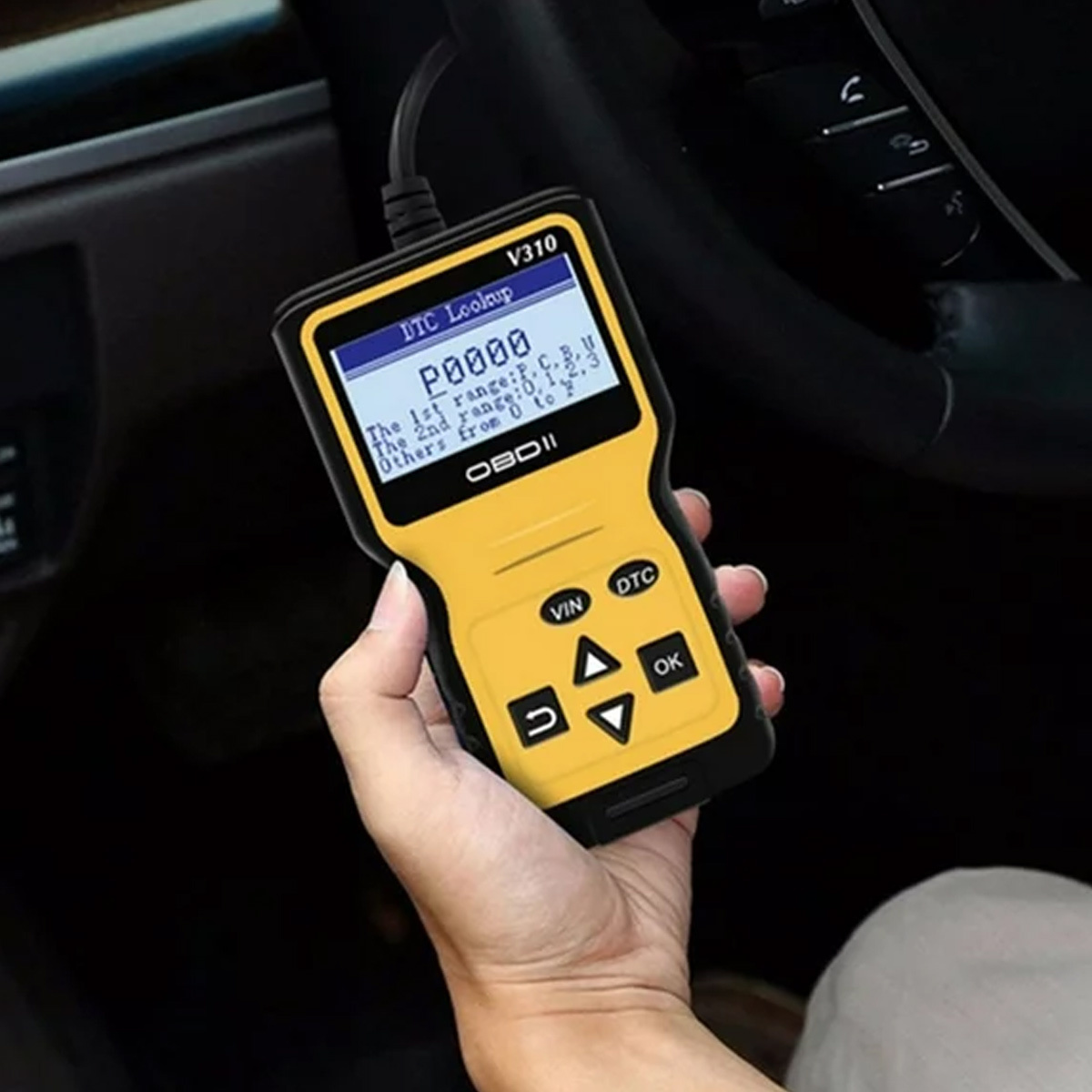 Eine Hand hält einen gelb-schwarzen OBDII-Scanner, der einen Code anzeigt. Der Scanner befindet sich in einem Auto mit einem Lenkrad im Hintergrund.