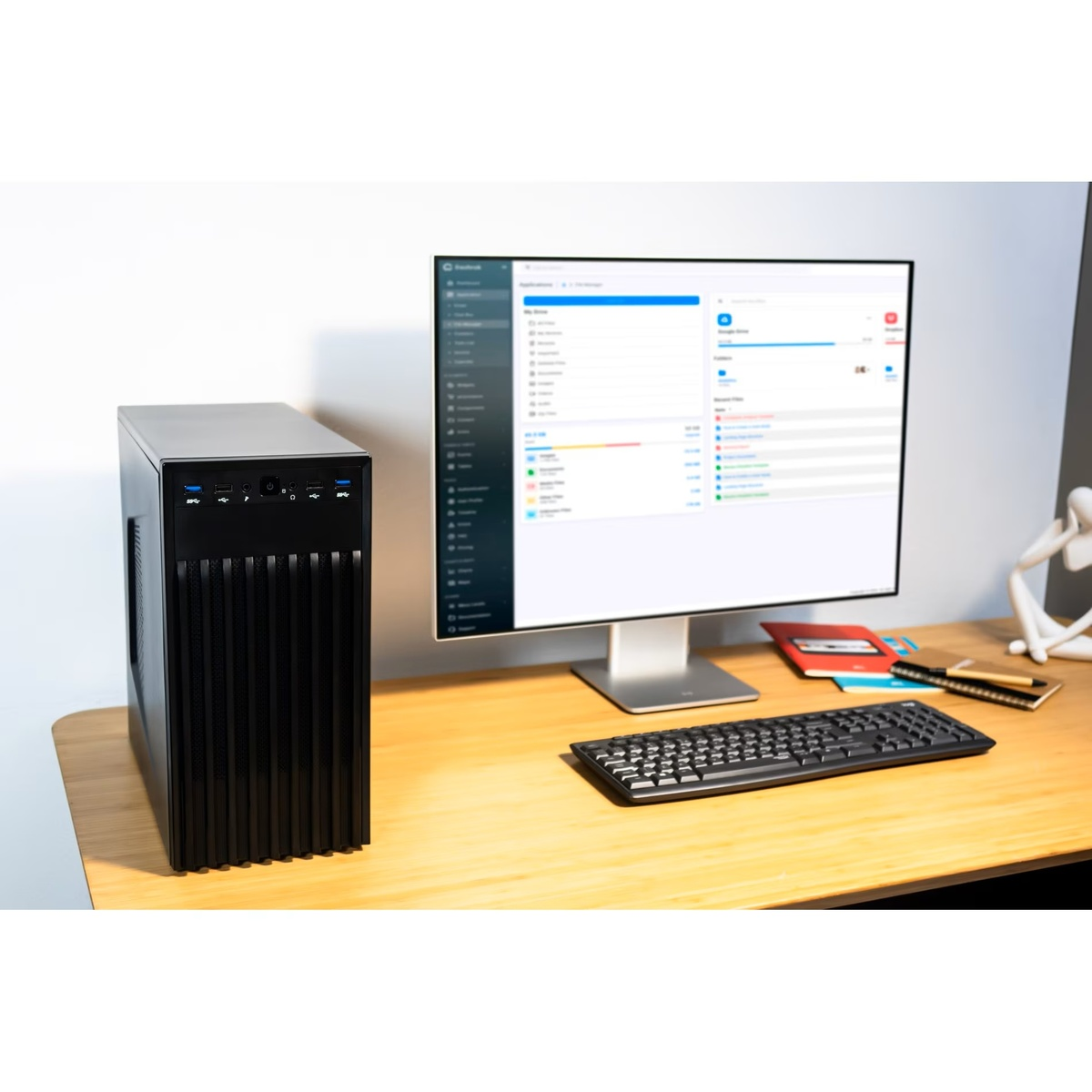 Czarna wieża komputera stoi obok monitora wyświetlającego interfejs zarządzania plikami, klawiaturę i notatnik na drewnianym biurku.