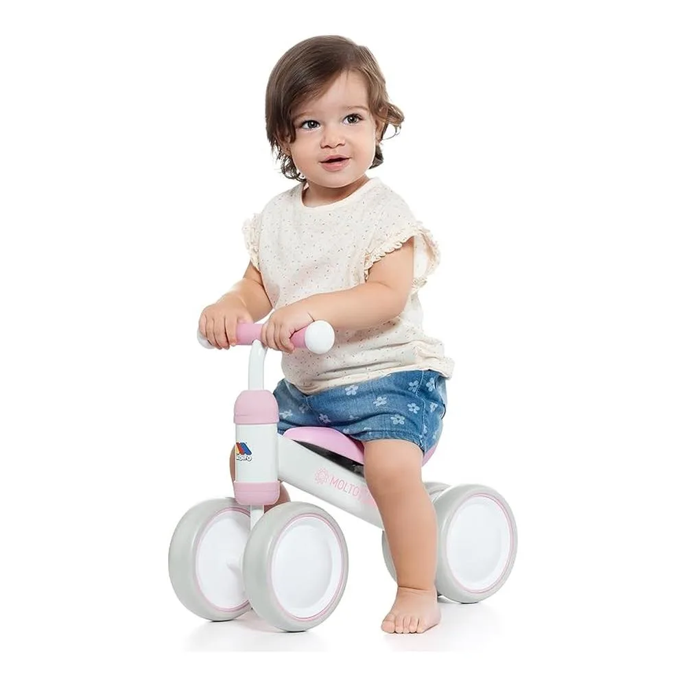 Un niño pequeño está sentado en una bicicleta de equilibrio rosa y blanca. Viste camisa blanca y pantalones cortos azules, sonríe y mira hacia adelante.