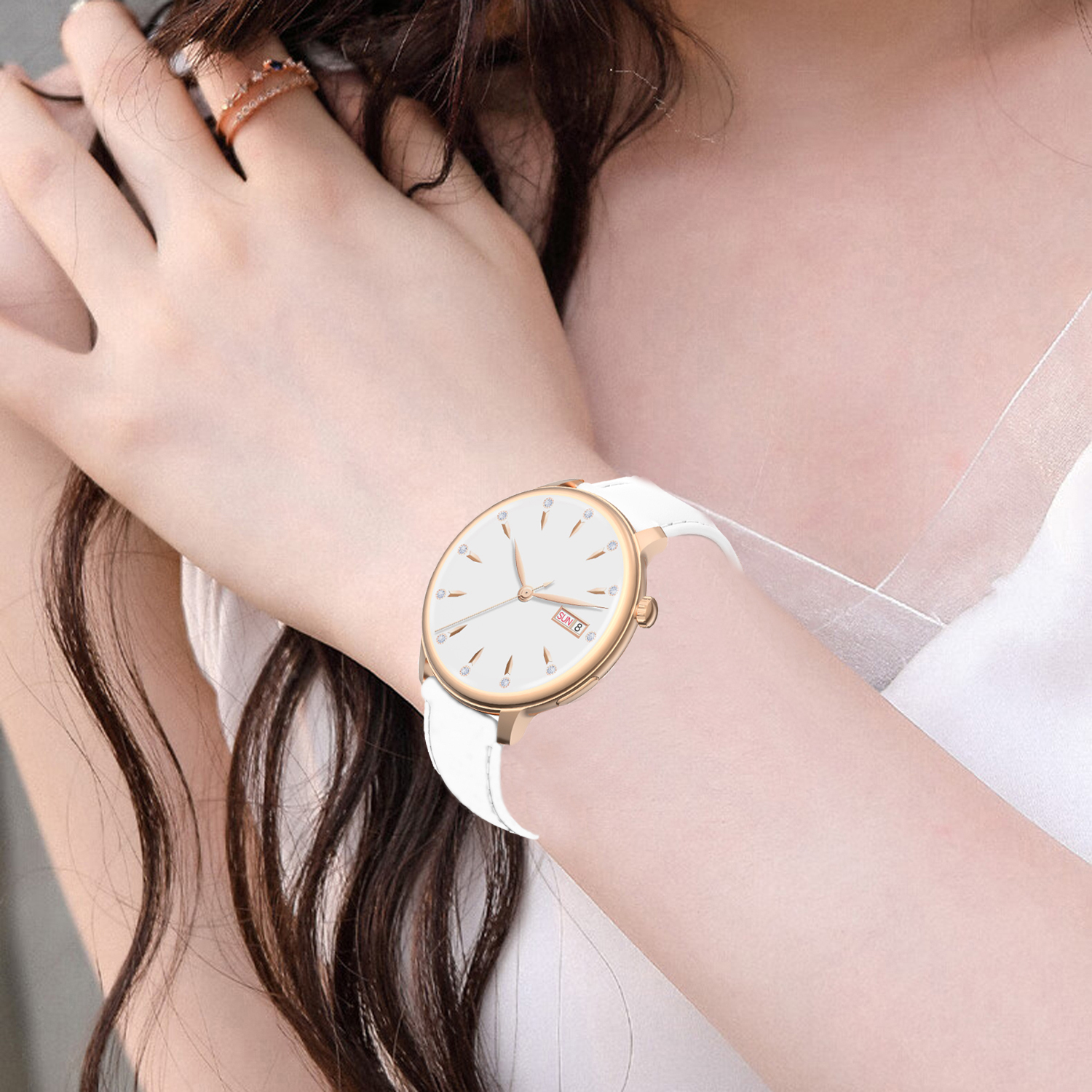A woman's wrist with a gold watch with a white face and band. A ring is on the adjacent finger. The woman wears a white top.
