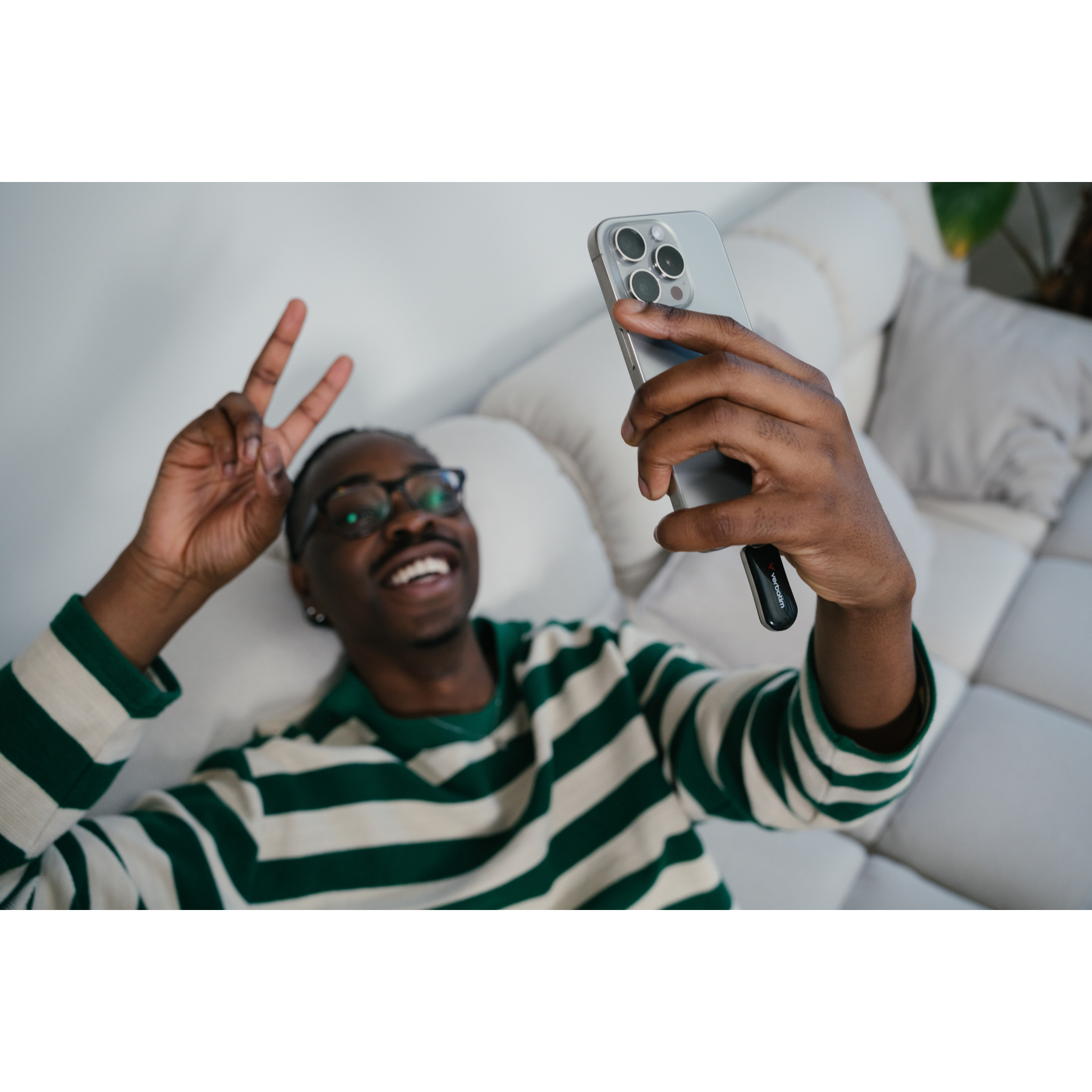 Mężczyzna robi sobie selfie, trzymając telefon w jednej ręce, robiąc znak pokoju drugą, uśmiechając się. Zielono-biała koszula w paski.