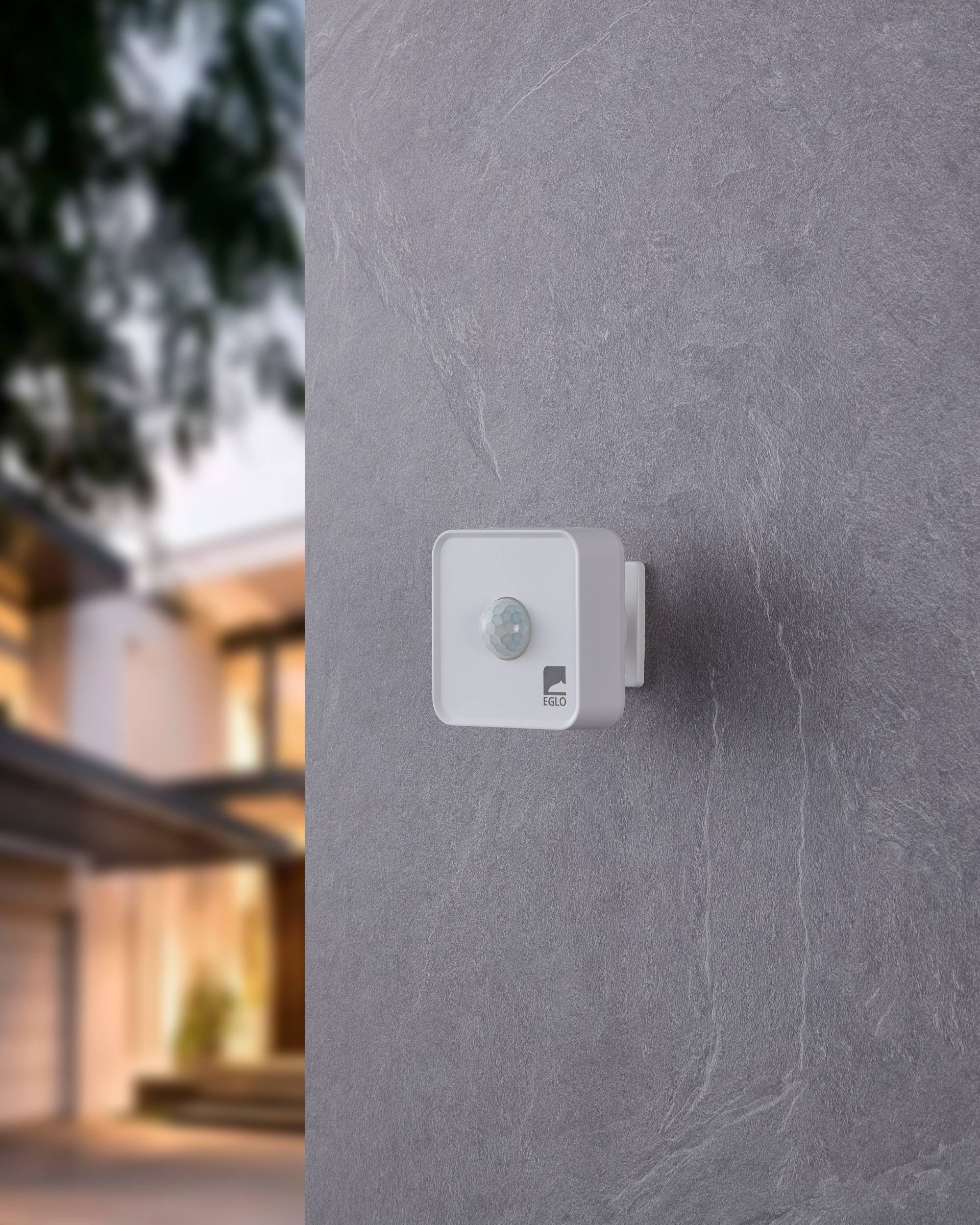 Sensore di movimento bianco su muro di pietra grigia. Casa sfocata sullo sfondo. Logo EGLO visibile sul sensore.