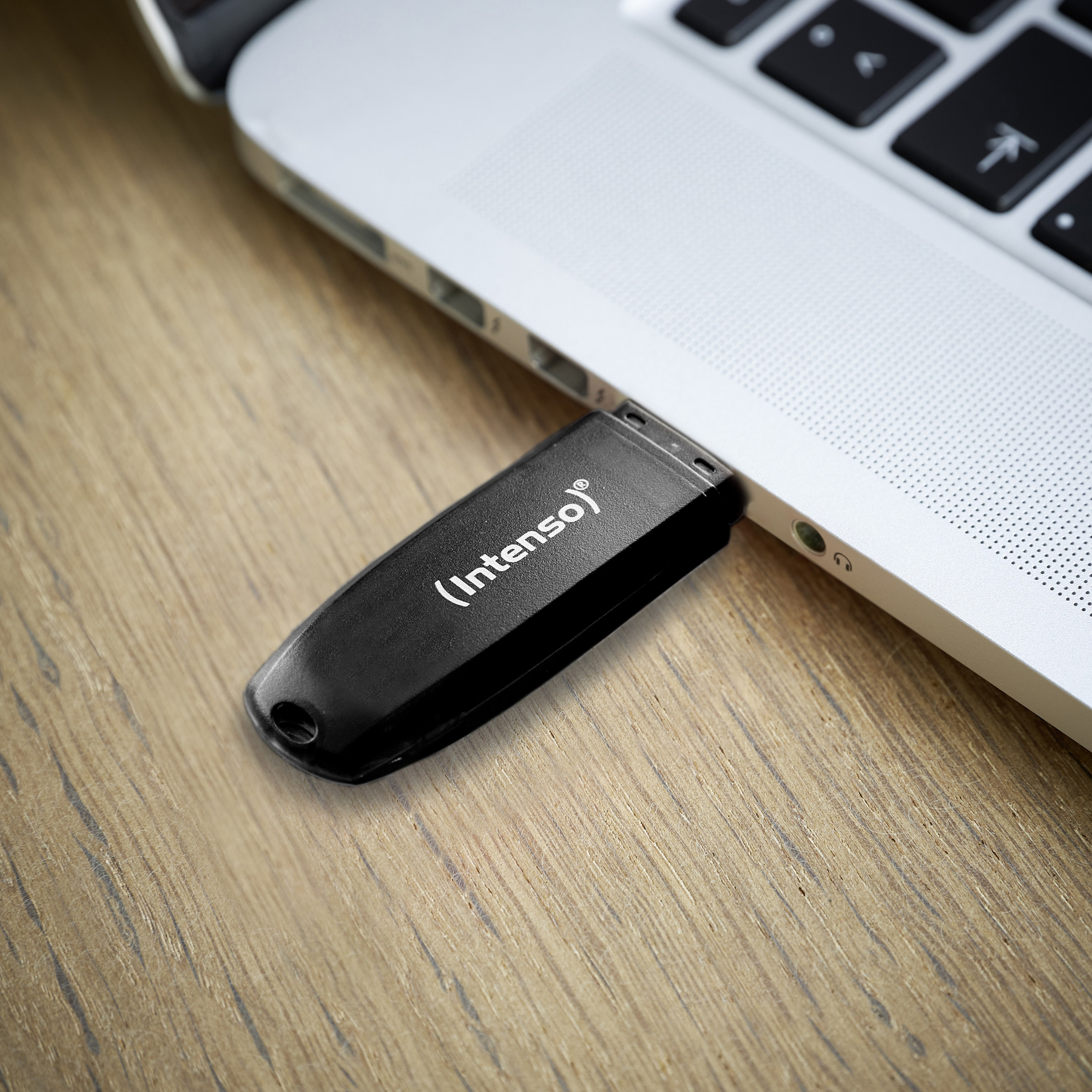 Ein schwarzer USB-Stick mit der Aufschrift 'Intenso' ist an einen silbernen Laptop angeschlossen. Der Laptop steht auf einer Holzoberfläche.