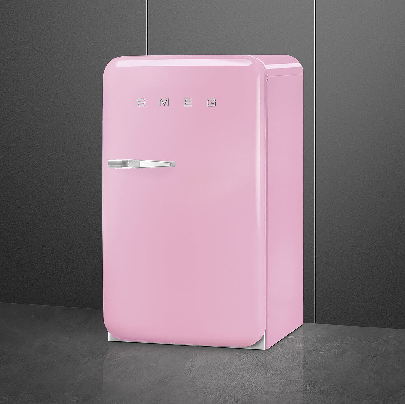 Un refrigerador retro SMEG rosa. Tiene un asa plateada y el nombre de la marca en la parte superior. Se encuentra sobre una superficie gris contra una pared gris oscura.
