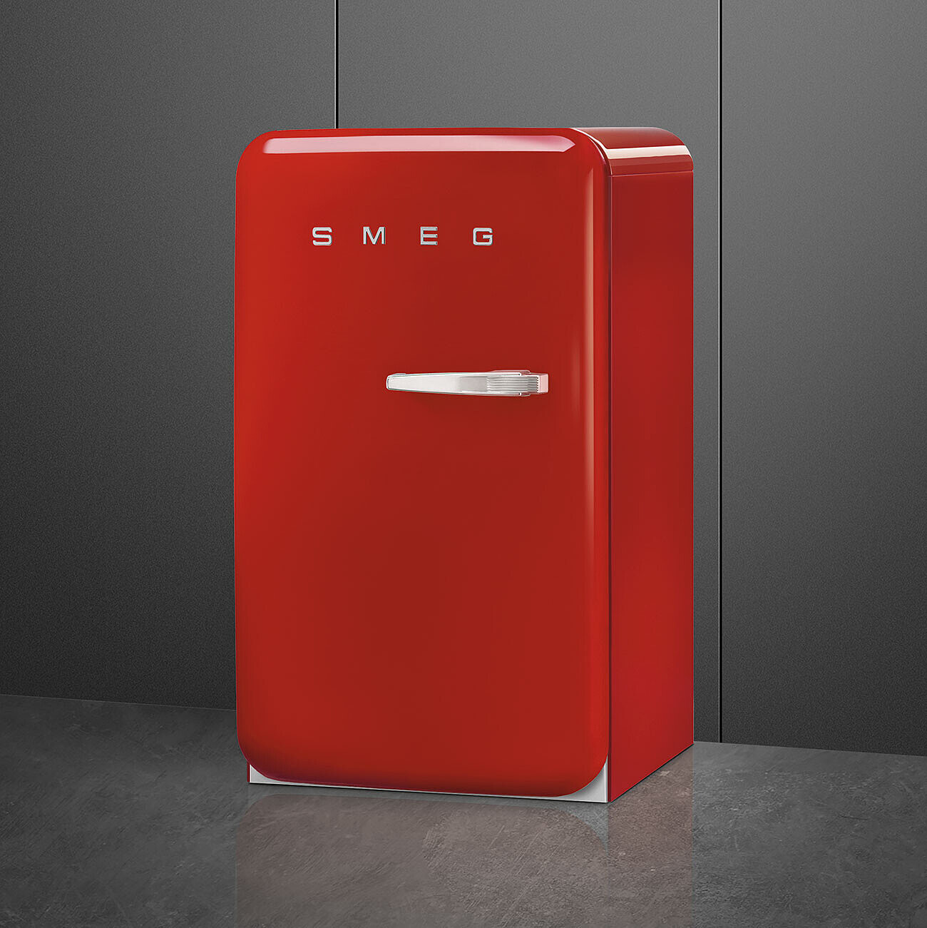 Un refrigerador SMEG rojo está sobre un piso gris contra una pared gris oscura. Tiene una manija plateada y el nombre de la marca.
