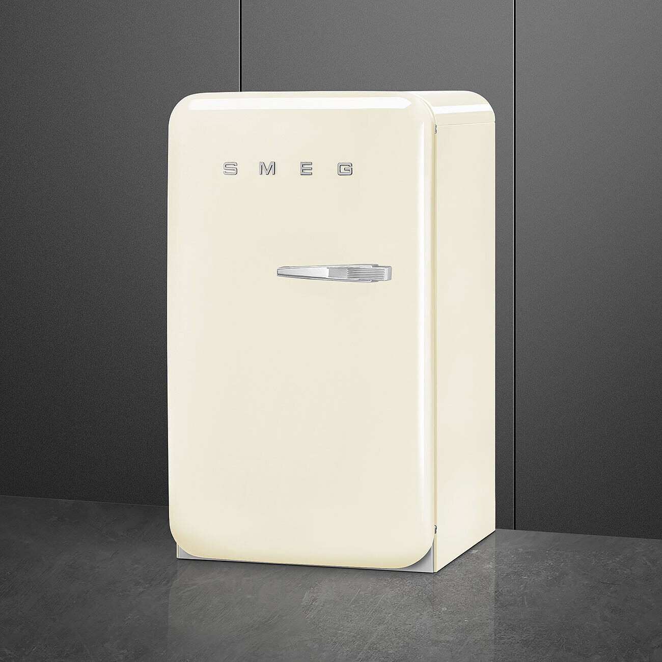 Un refrigerador SMEG color crema se encuentra sobre una superficie gris contra una pared gris oscuro. El mango es cromado y la marca es visible.
