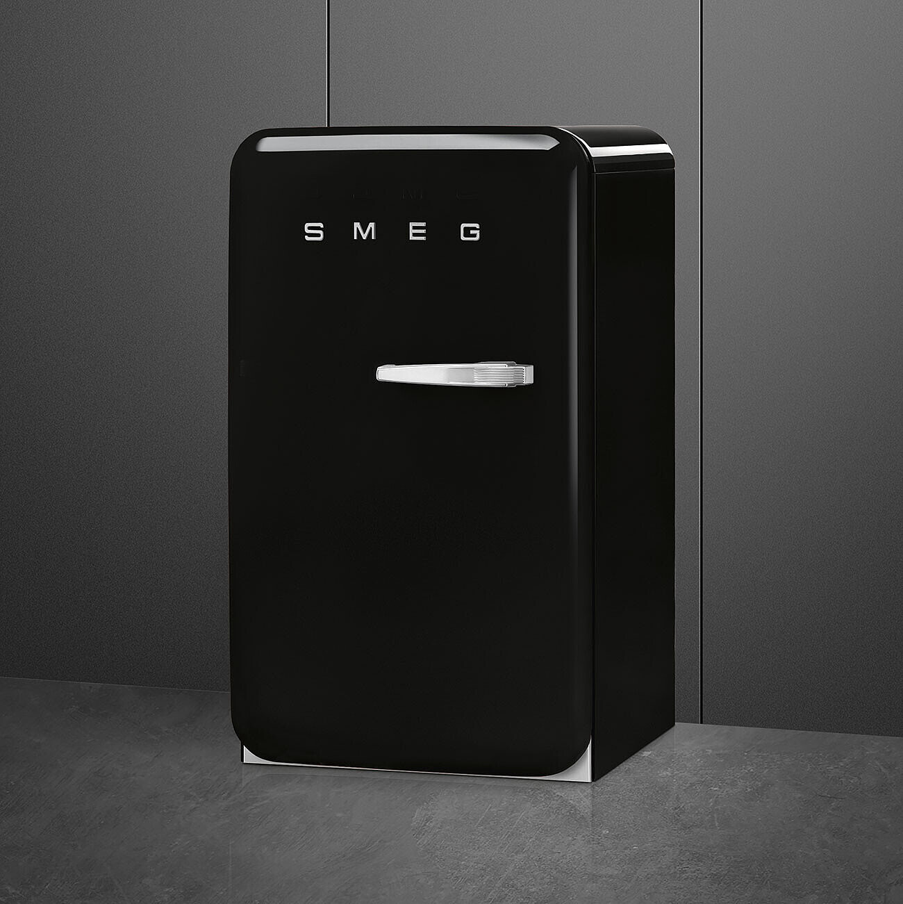 Un refrigerador SMEG negro está sobre un piso gris contra una pared gris. Tiene una manija plateada y el logotipo de SMEG.