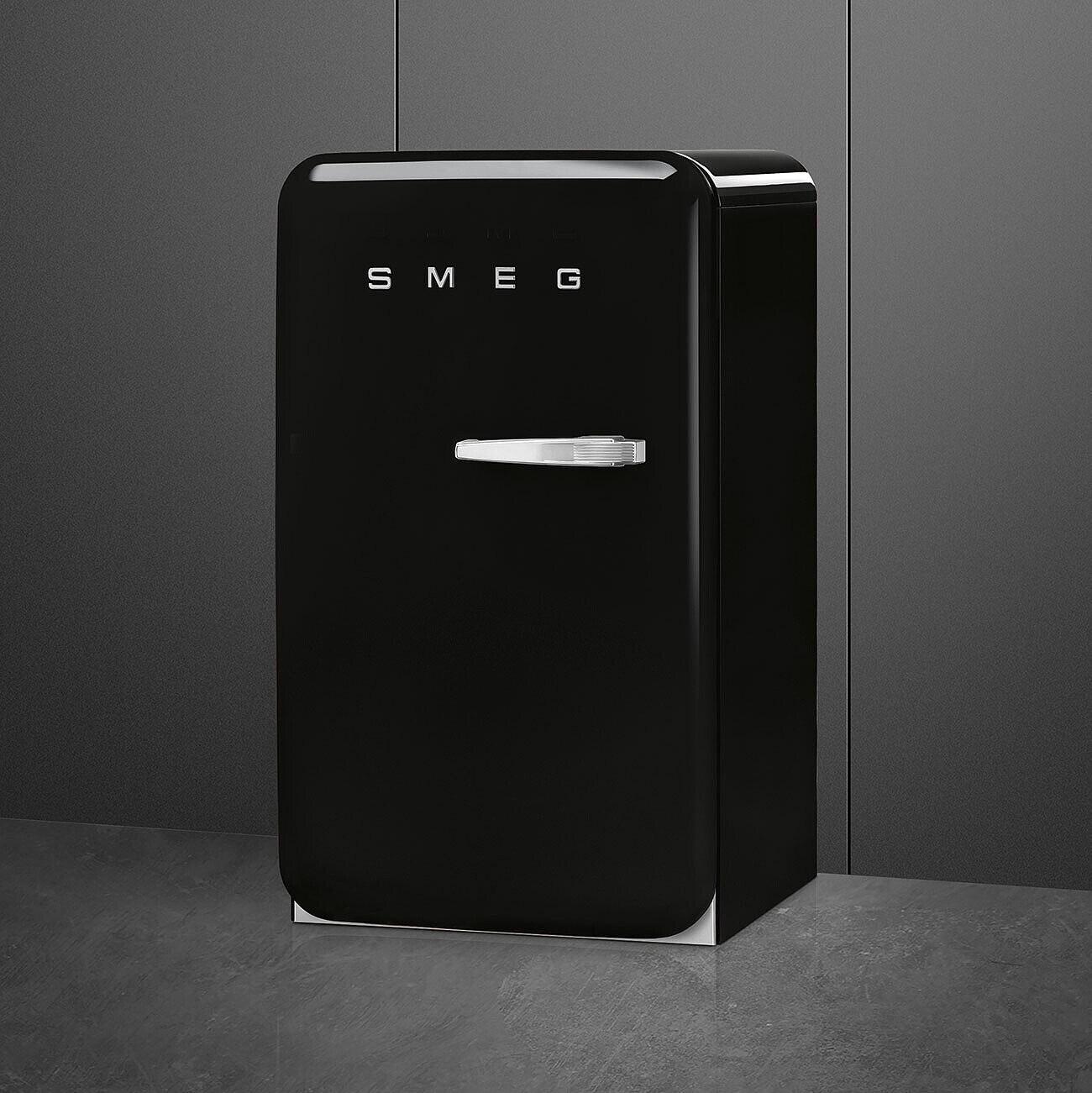 Un refrigerador SMEG negro se encuentra contra una pared gris oscura. Tiene una manija plateada y la palabra 'SMEG' al frente.