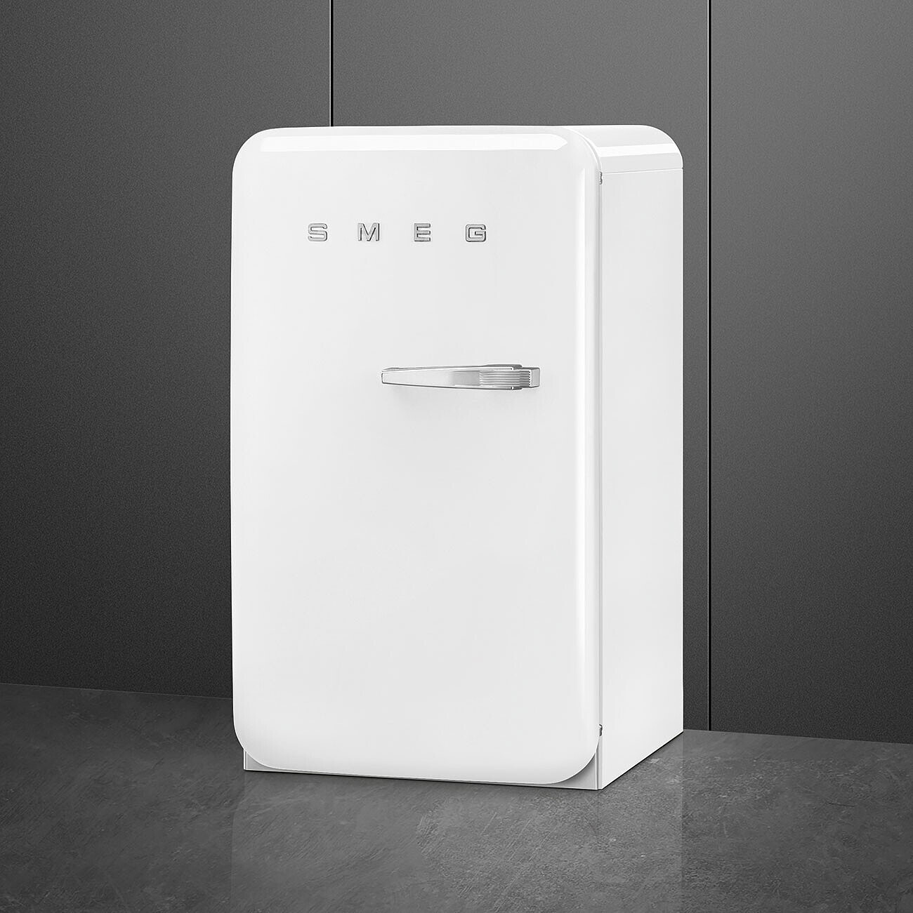 Un refrigerador SMEG blanco está contra una pared gris. Tiene un asa plateada y el logotipo SMEG en la puerta.