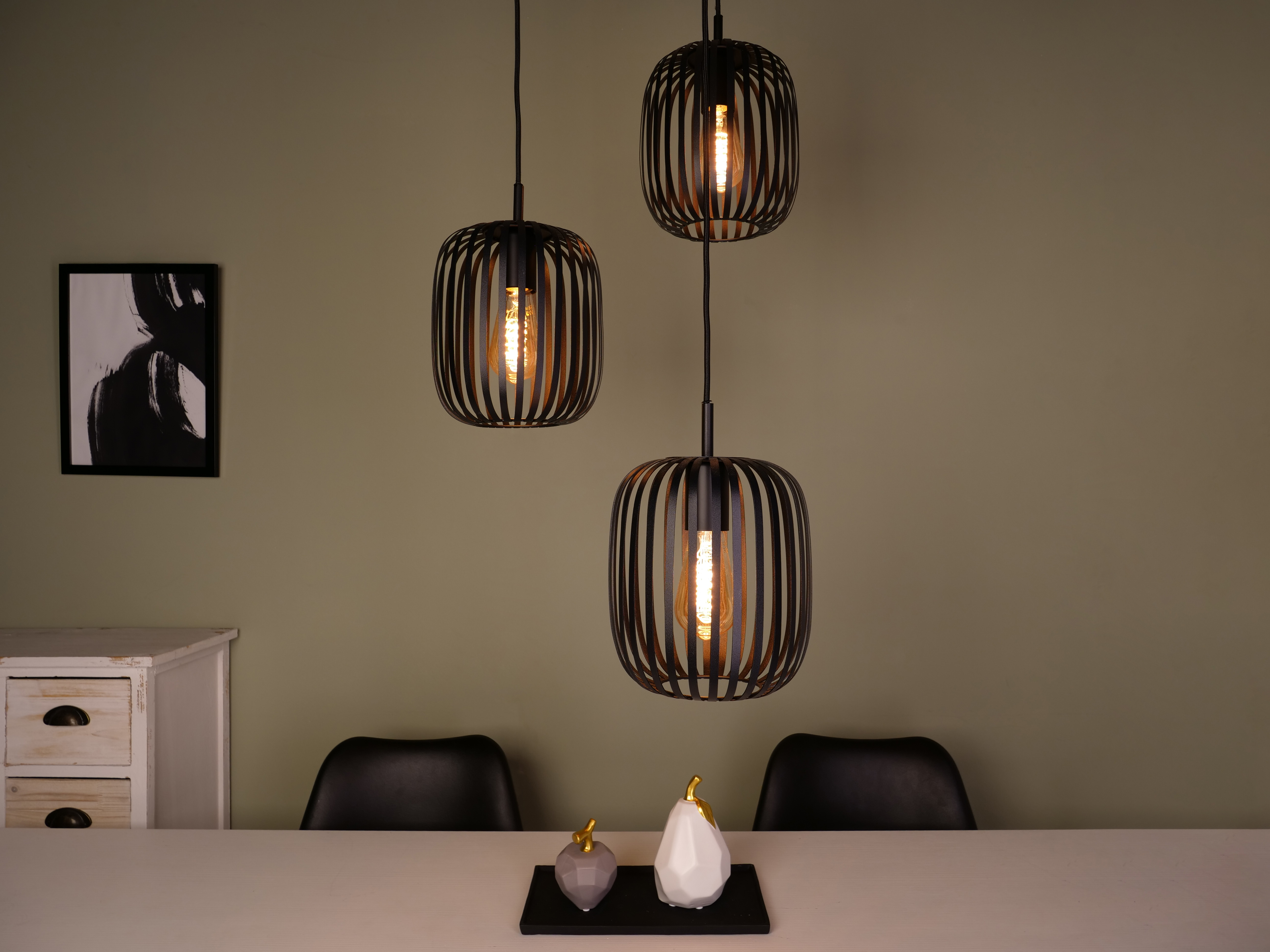 Drie zwarte hanglampen met Edison-lampen hangen boven een tafel met decoratieve objecten. Een zwart-wit kunstwerk hangt aan de muur.
