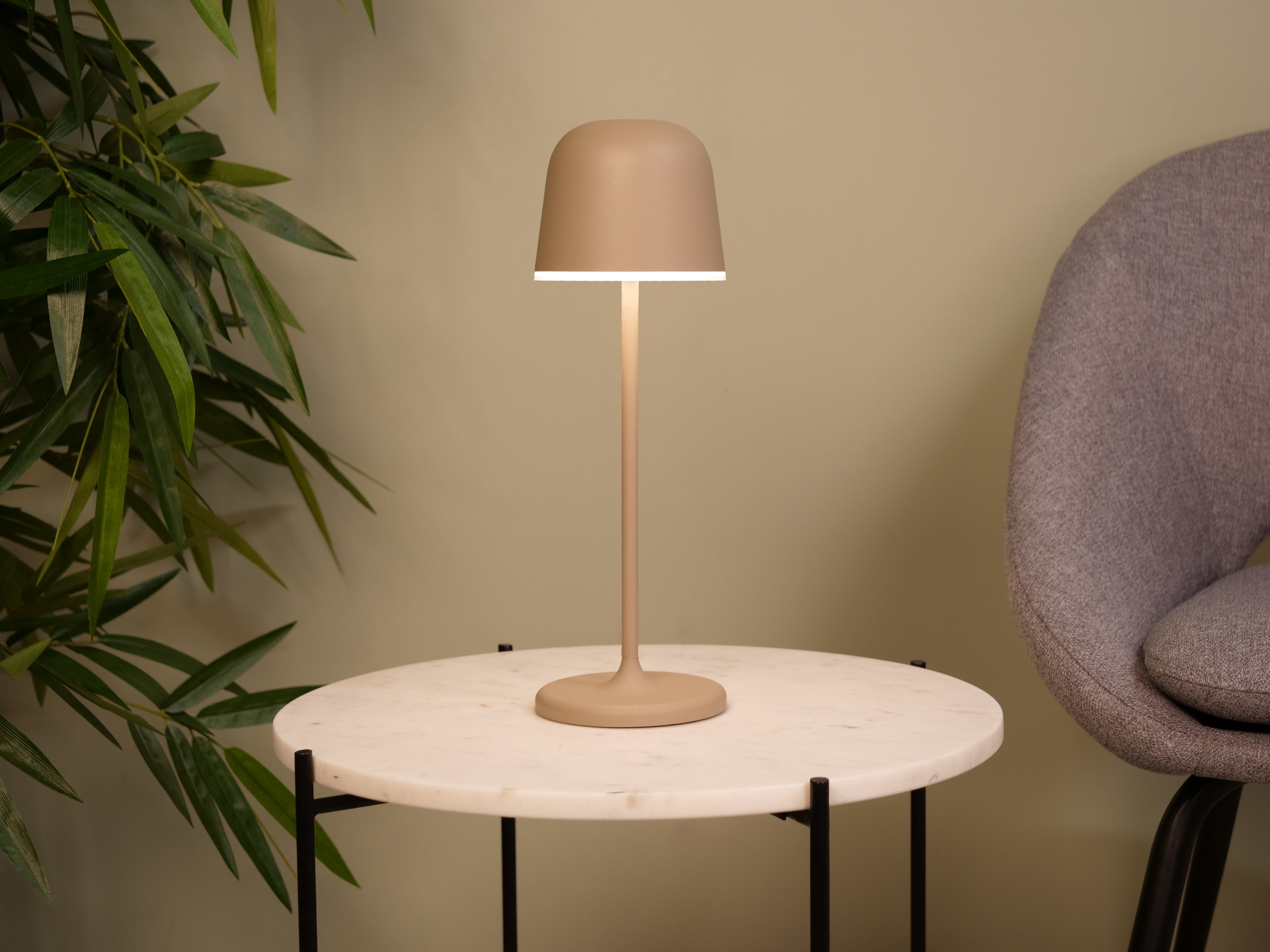 Eine beige Lampe auf einem Marmortisch, Bambusblätter links, grauer Stuhl im Hintergrund.
