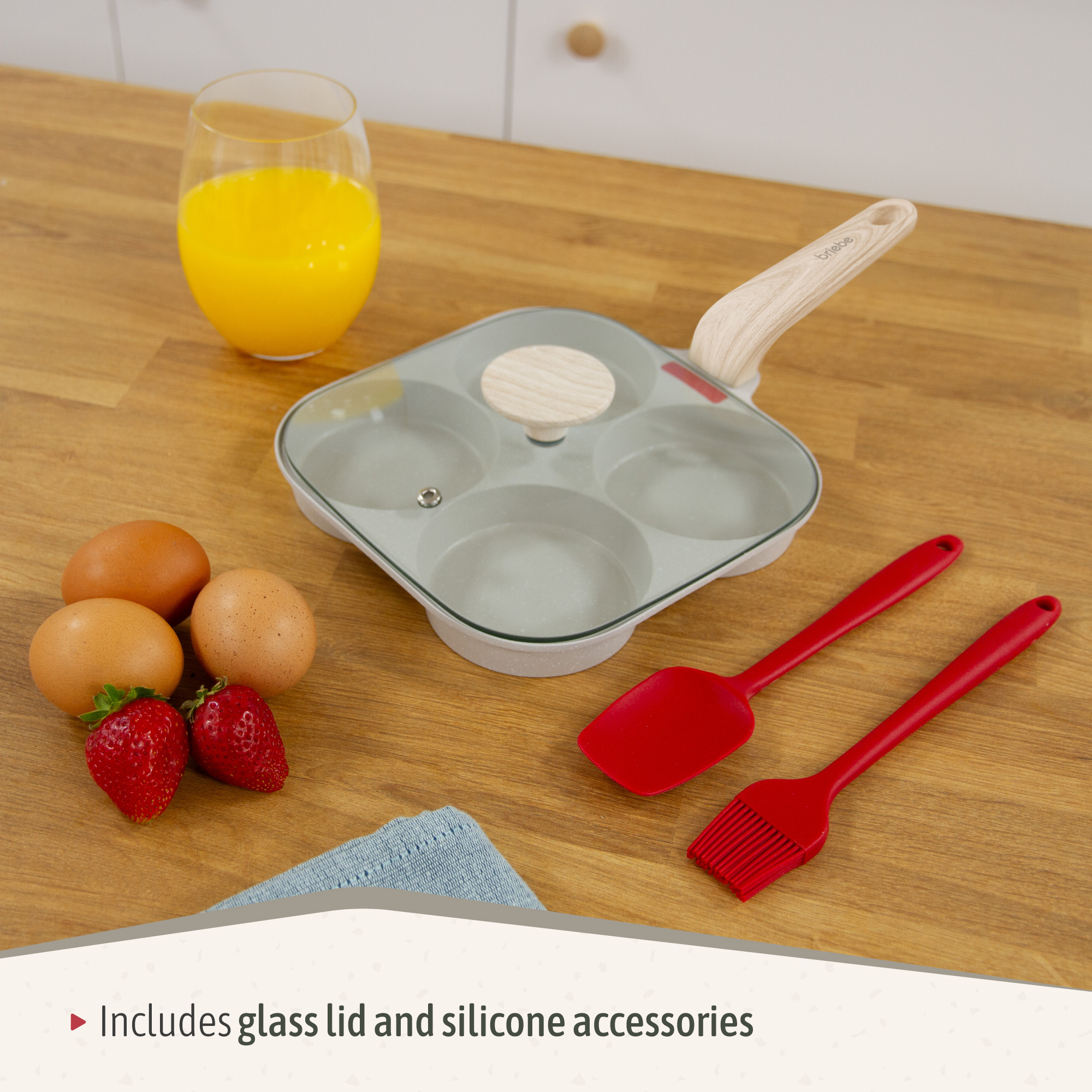 Una sartén con tapa, huevos, fresas, jugo de naranja y utensilios rojos sobre una superficie de madera. Incluye tapa de vidrio y accesorios de silicona.