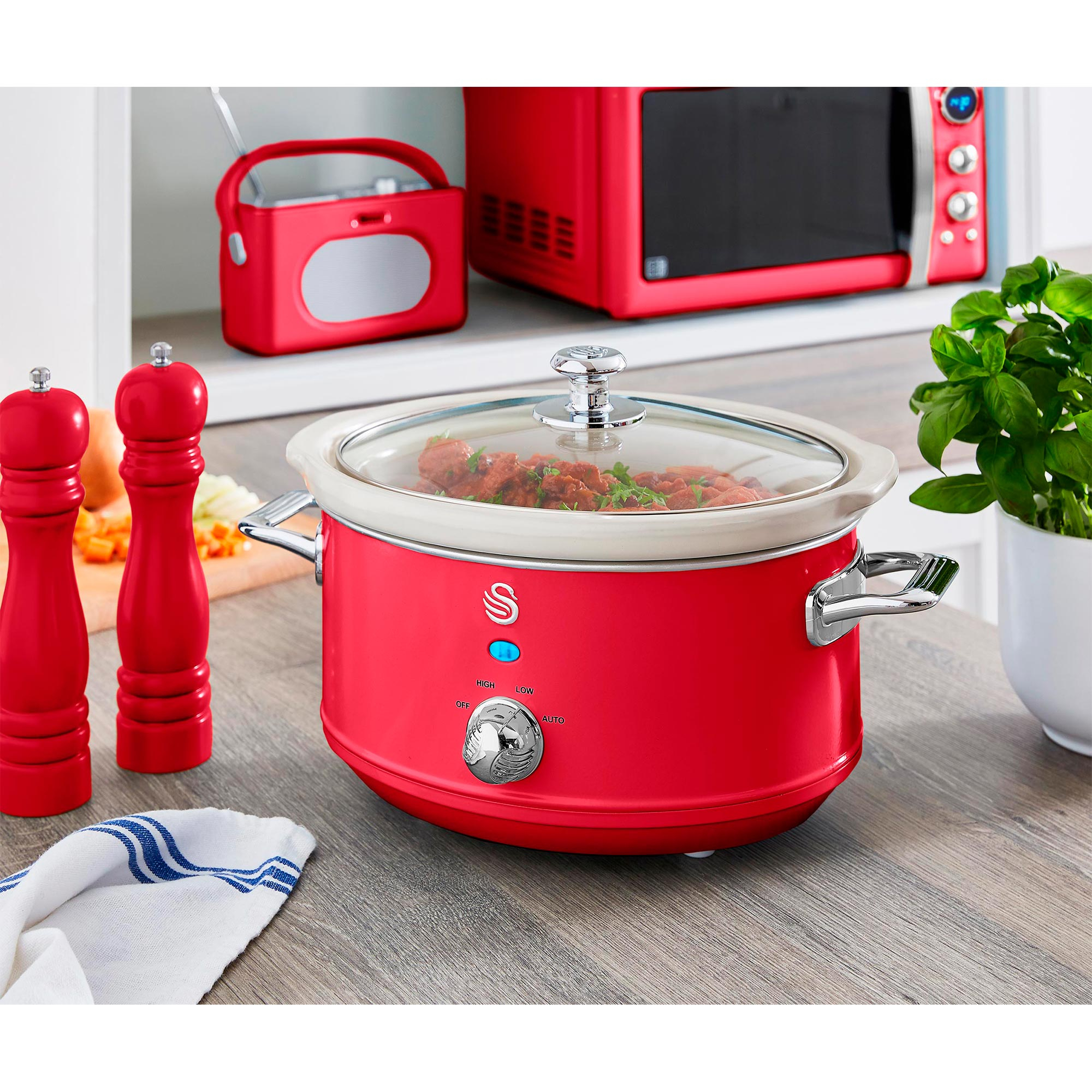 Ein roter Slowcooker steht auf einer Theke. Eine Mikrowelle, ein Radio und eine Basilikumpflanze befinden sich im Hintergrund.