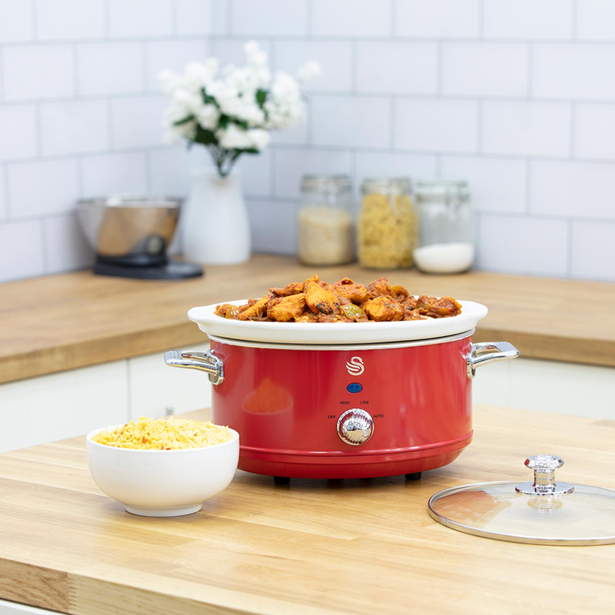 Ein roter Slow Cooker mit Essen steht auf einer Holztheke. Eine Schüssel mit Reis und ein Glasdeckel sind in der Nähe. Weiße Fliesenwand im Hintergrund.