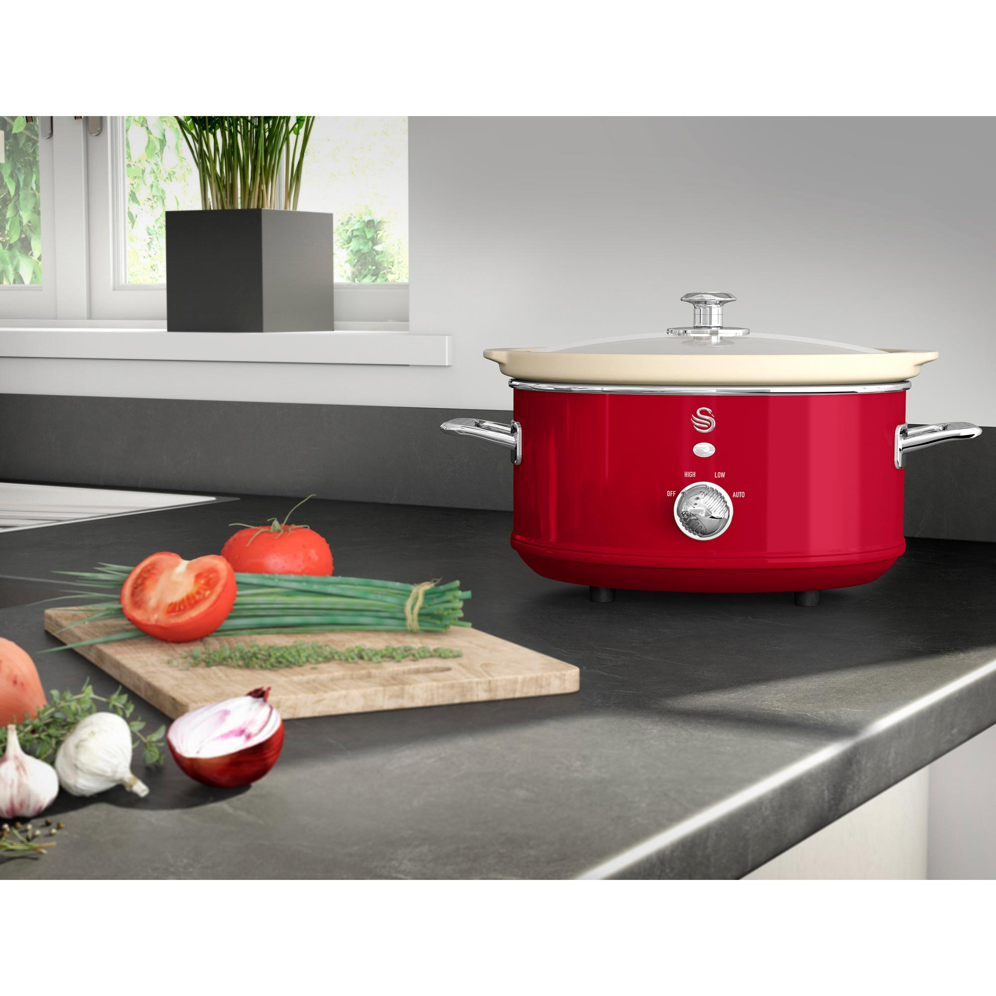 Ein roter Slowcooker mit Gemüse und Kräutern auf einem Schneidebrett steht auf einer Küchentheke.
