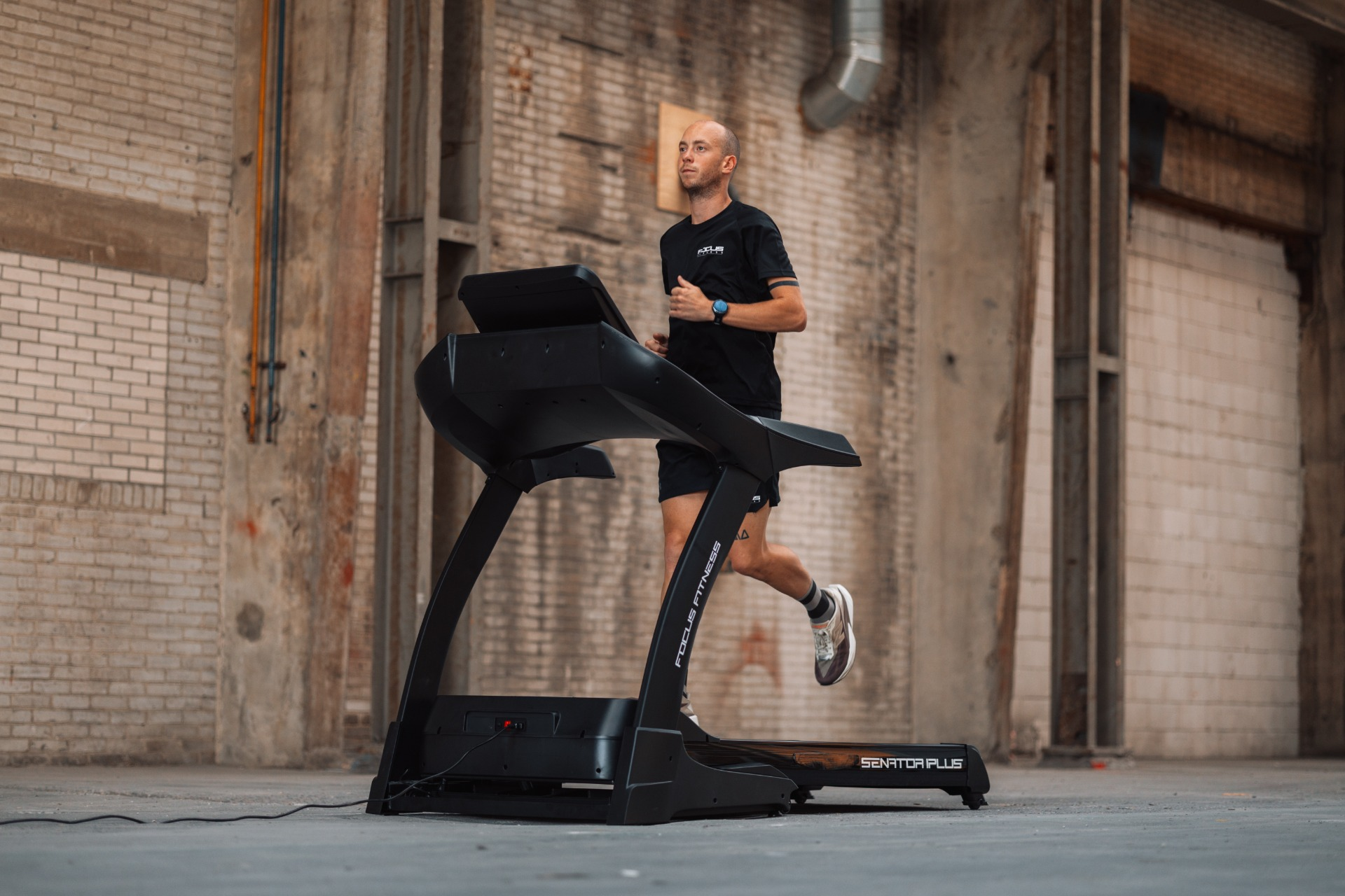 A man runs on a treadmill in an industrial space. The treadmill is black, and the man wears black workout clothes.