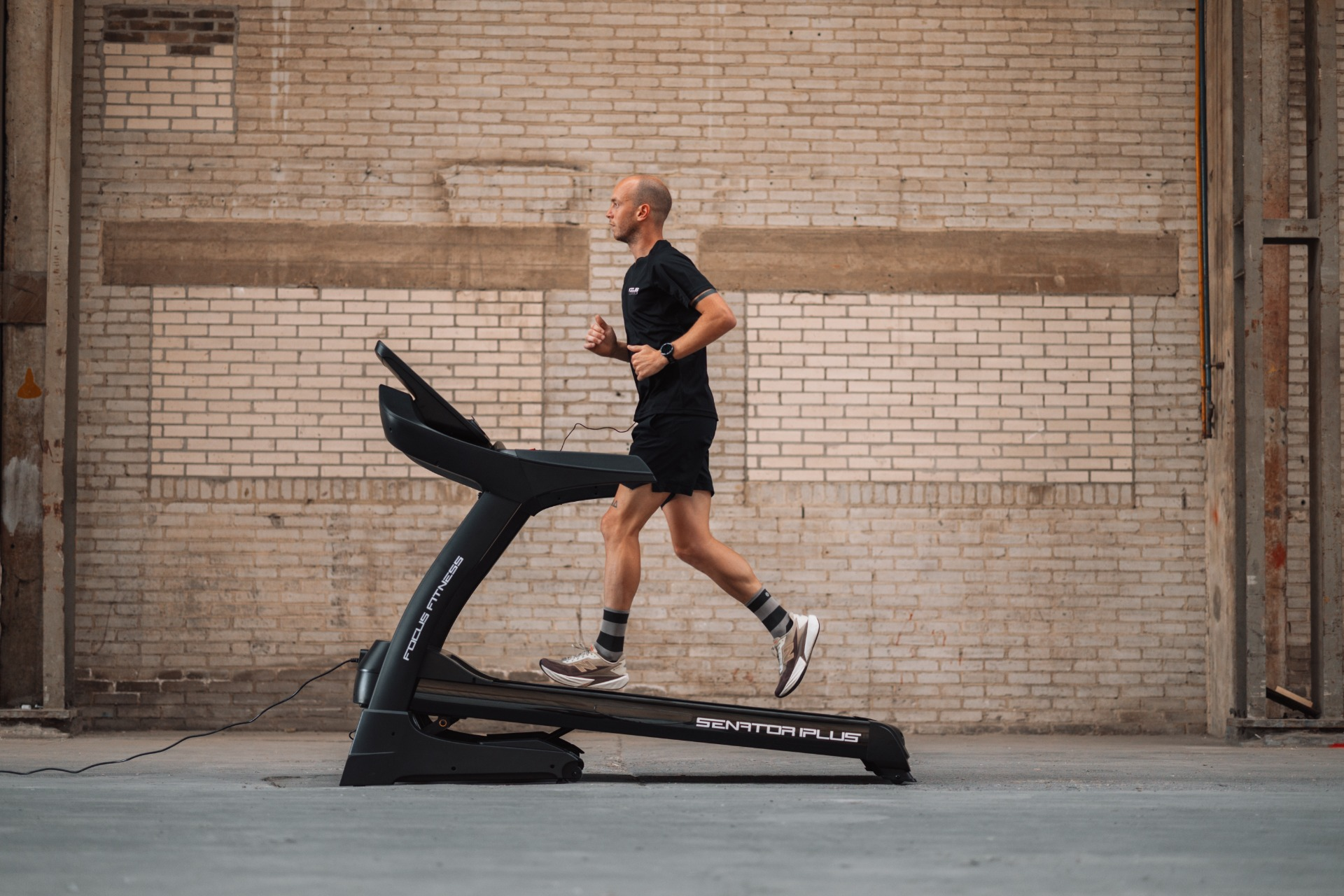 A man runs on a treadmill in a gym. He wears black shorts and shirt. A brick wall is in the background.