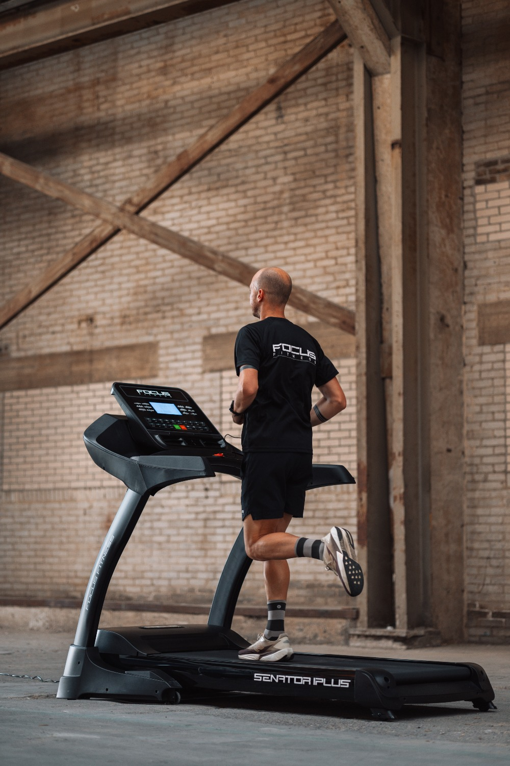 A man runs on a treadmill in a brick-walled room. He wears black shorts and a Focus Fitness shirt.