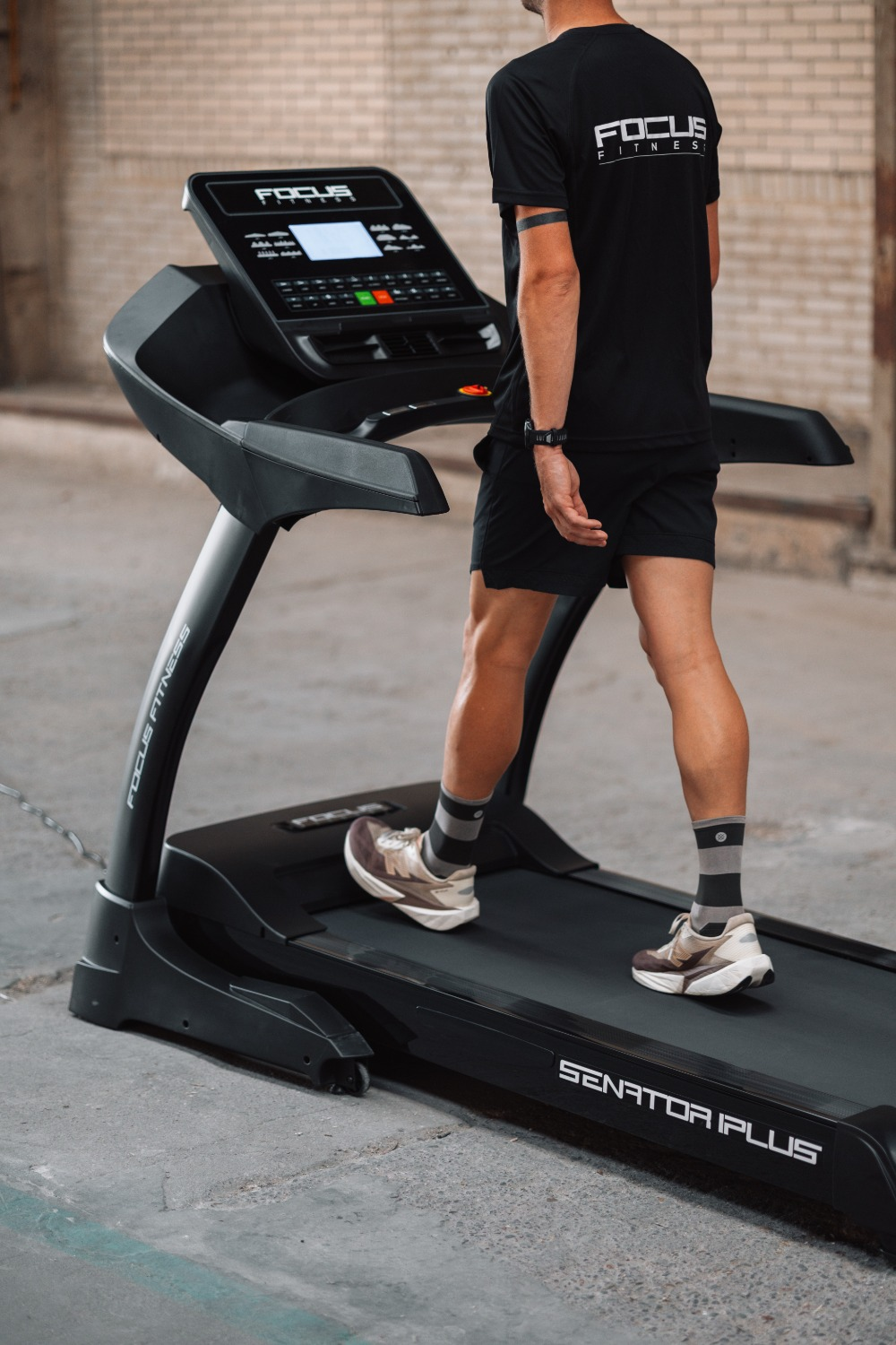 A man in black shorts and shirt walks on a black treadmill. The treadmill is in a room with brick and concrete walls.