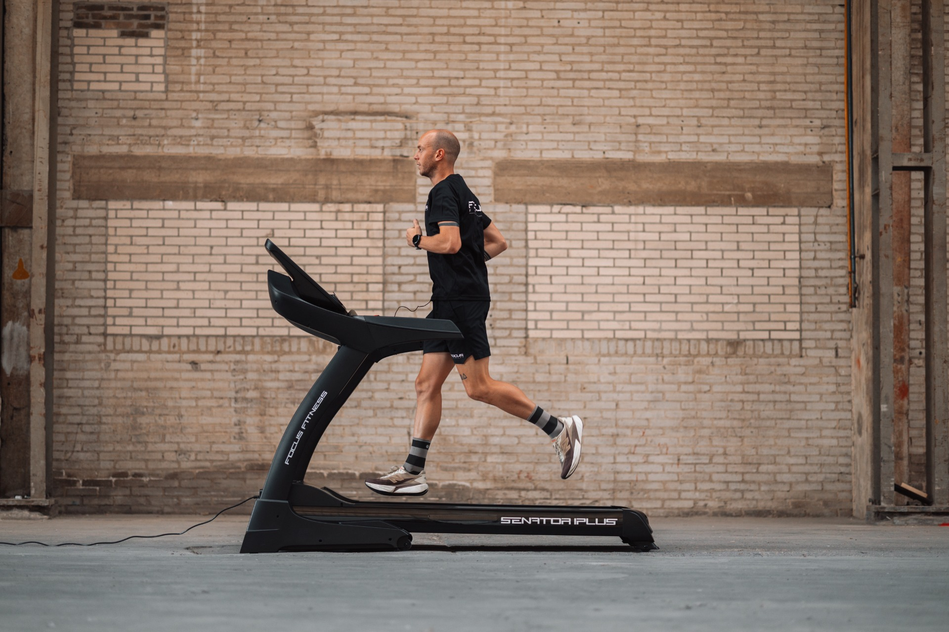 A man runs on a treadmill in front of a brick wall. He wears black shorts, shirt, and socks. The treadmill is black.