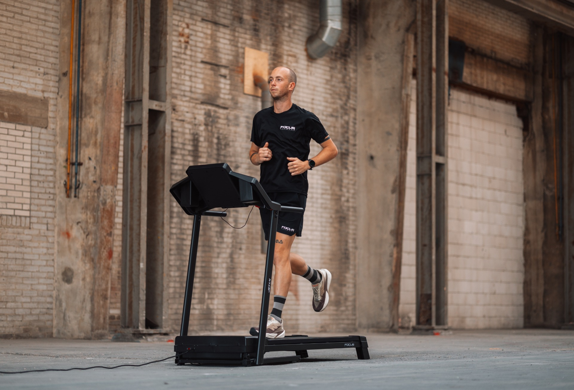 Man in black sportswear runs on a treadmill in an industrial setting. Background includes brick walls and metal structures.