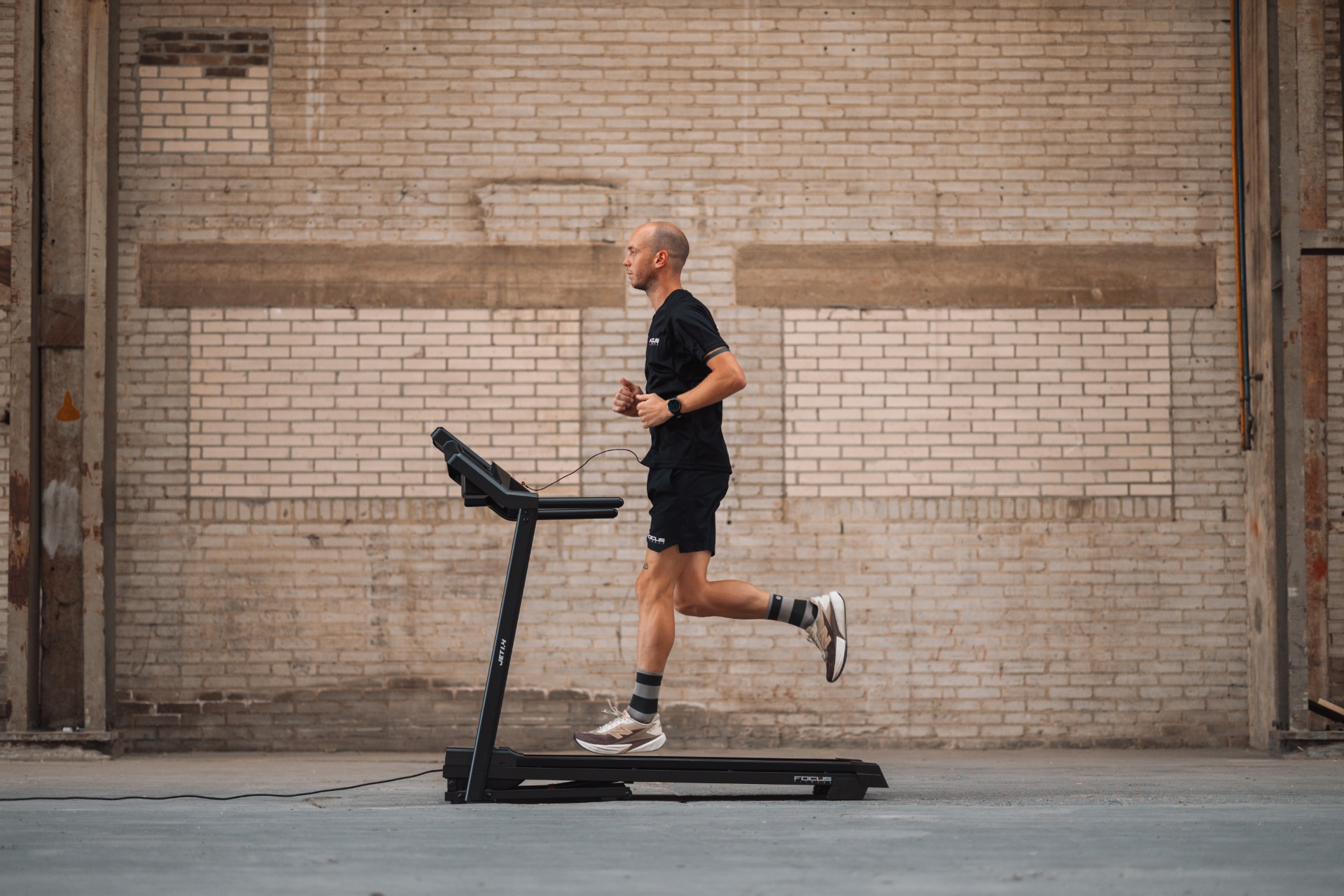 A man runs on a treadmill in front of a brick wall. He wears black shorts, a shirt, and running shoes.