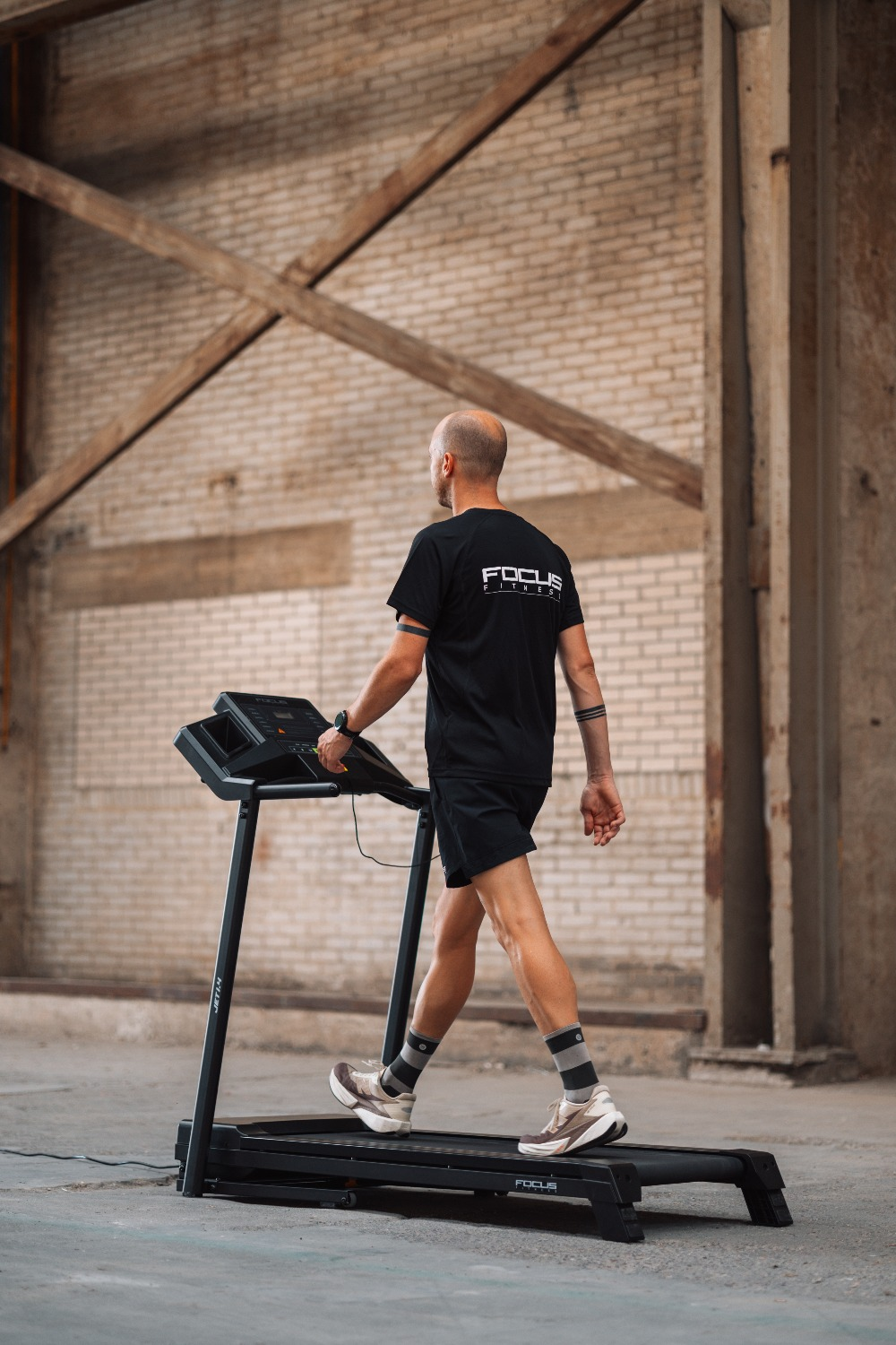 A man walks on a treadmill indoors. He wears black athletic wear. The background has brick and wooden beams.