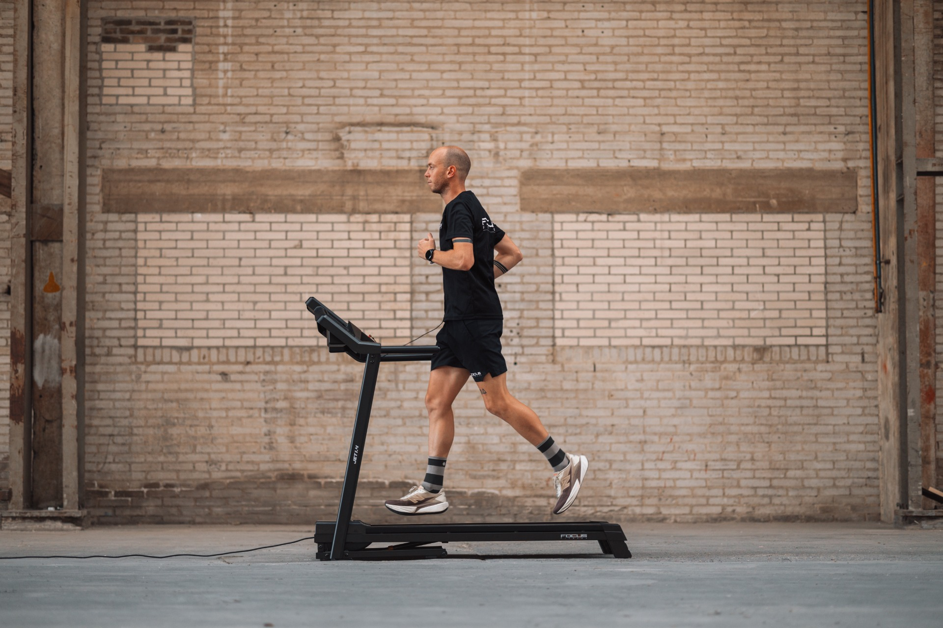 Man running on a treadmill in a warehouse. He wears black shorts, a t-shirt, and sneakers. The background is brick.