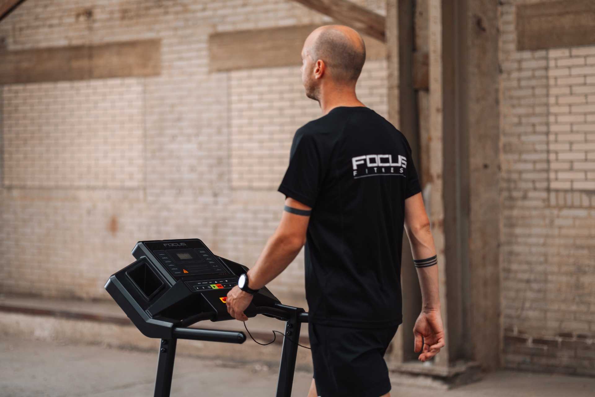 A man in black sportswear walks next to a treadmill. The background shows a brick wall and a concrete structure.