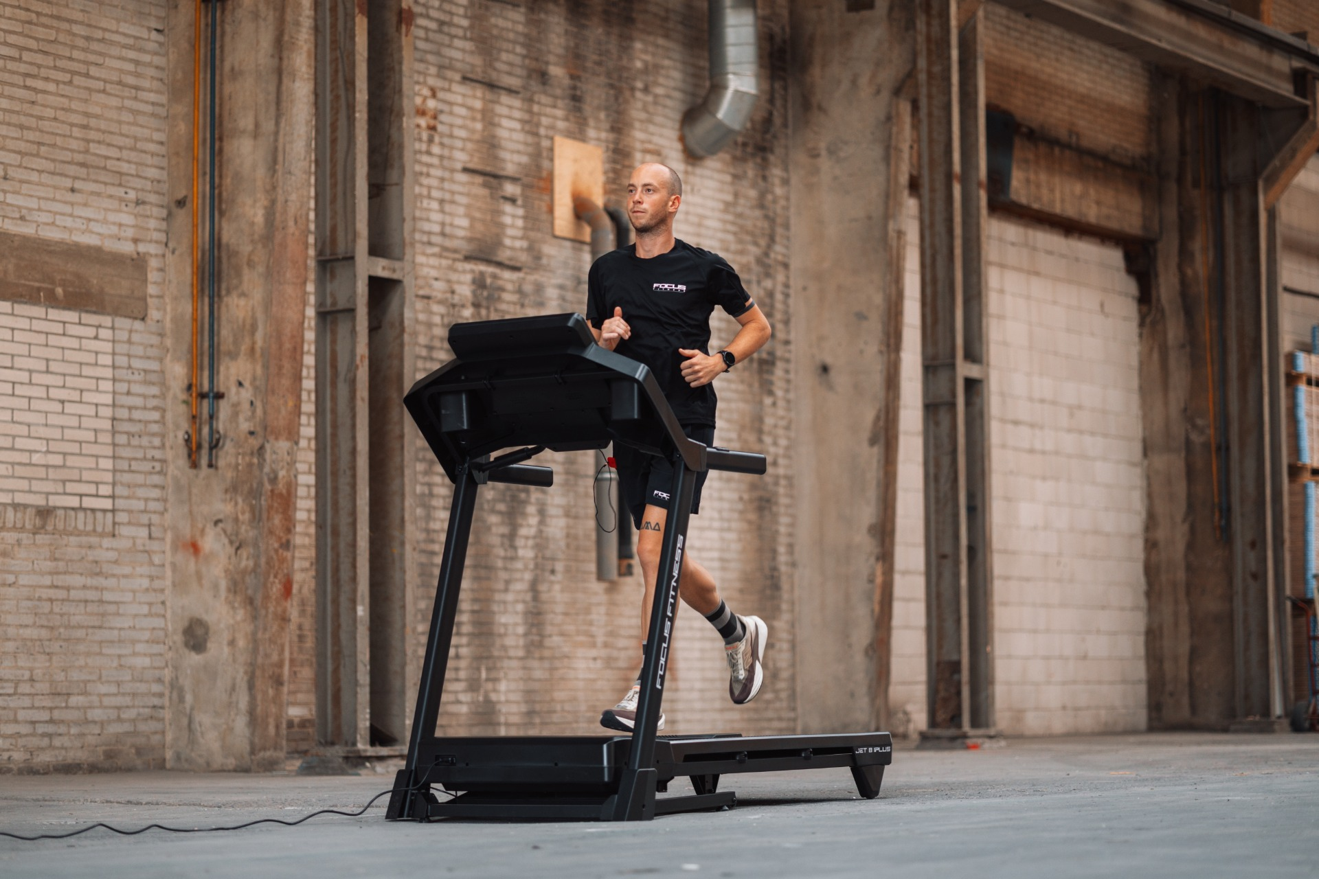 A man runs on a treadmill in an industrial setting. He wears black athletic clothes. The background is brick and concrete.