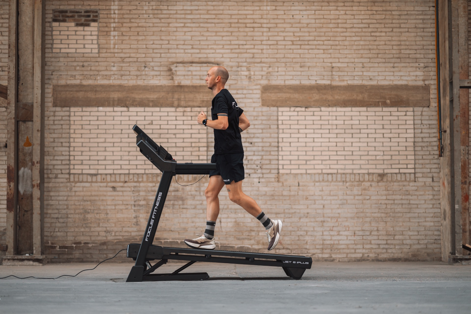 A man runs on a treadmill. He wears black shorts and shirt. The background is a brick wall.